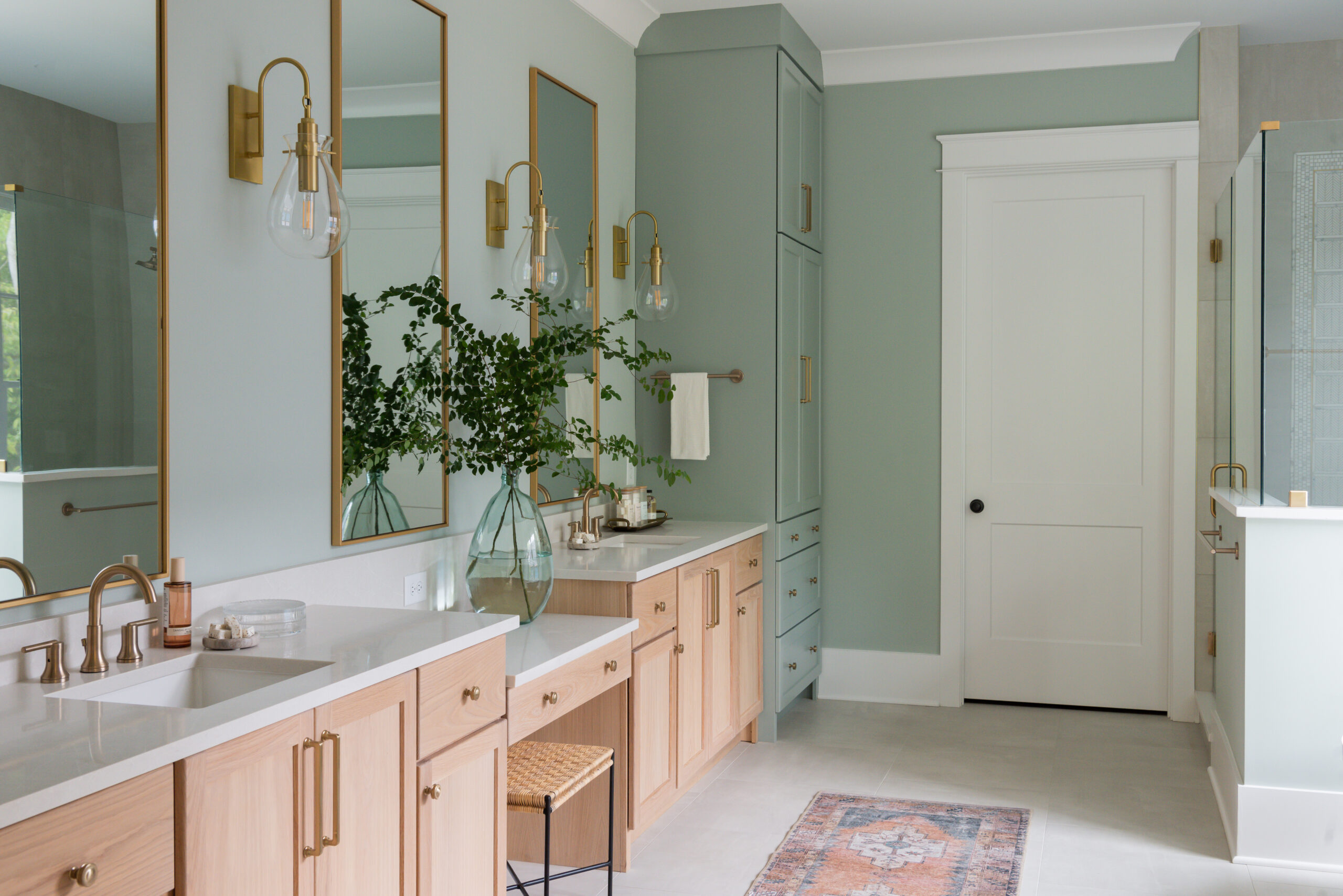 Dual light wood vanity with white countertops and a built in vanity seat with sage green walls and gold rimmed mirrors