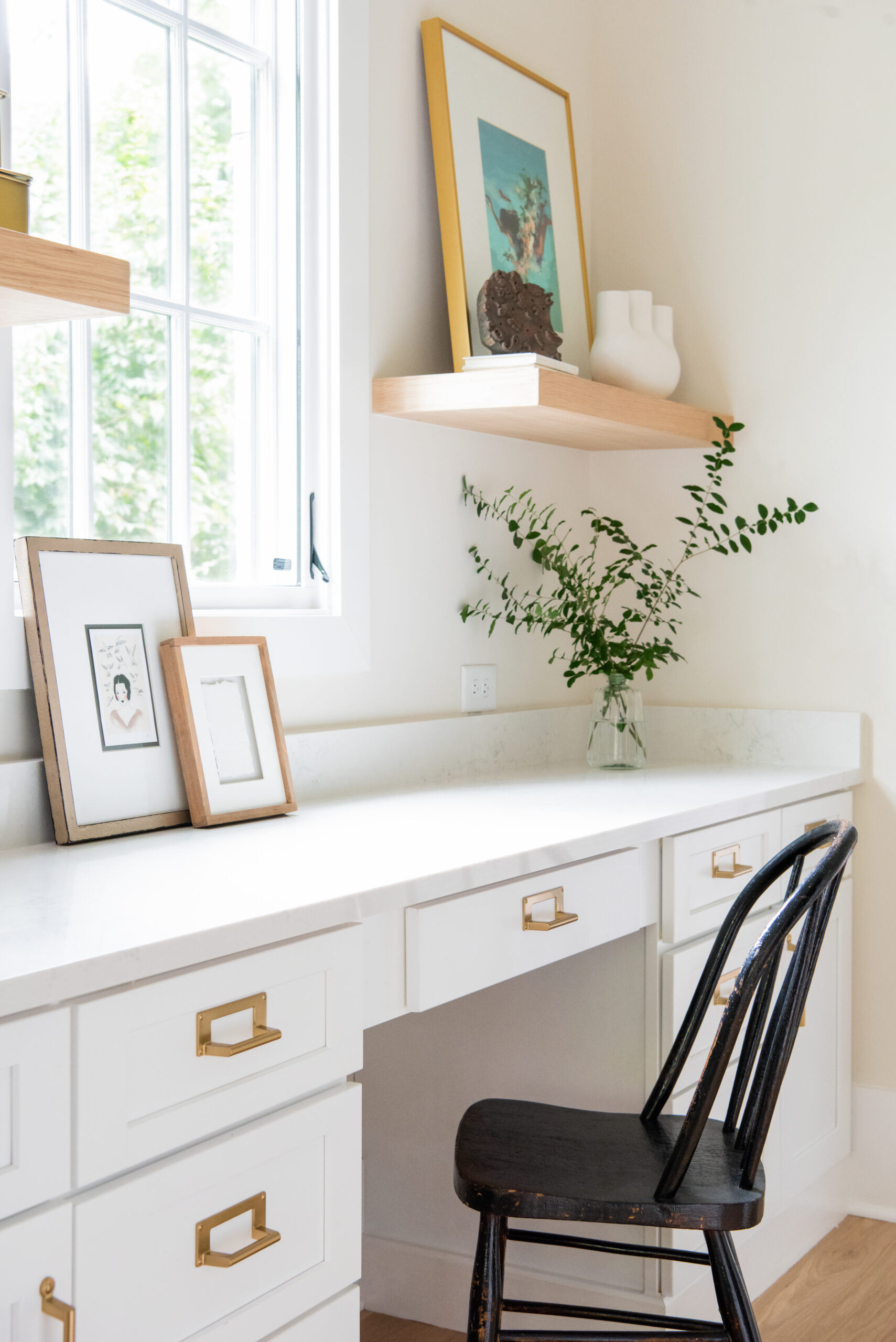 Office nook with white desk drawers and countertop, with white walls and floating natural wood stained shelves on either side of window and a black chair