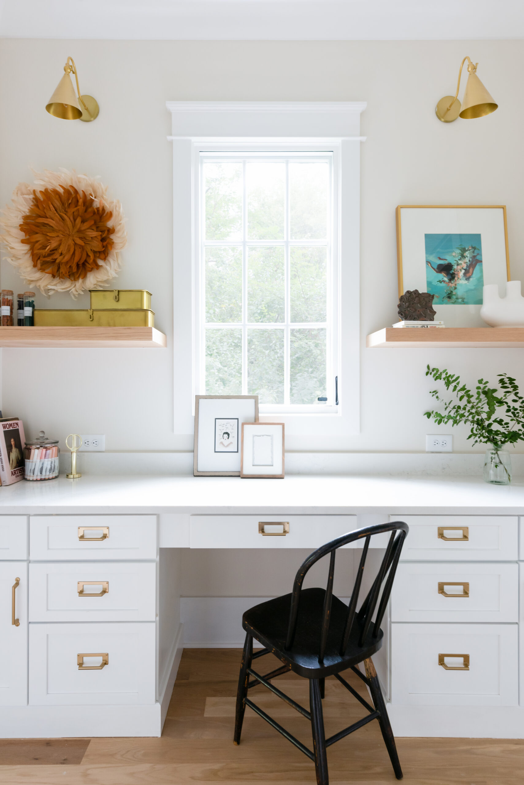 Office nook with white desk drawers and countertop, with white walls and floating natural wood stained shelves on either side of window and a black chair