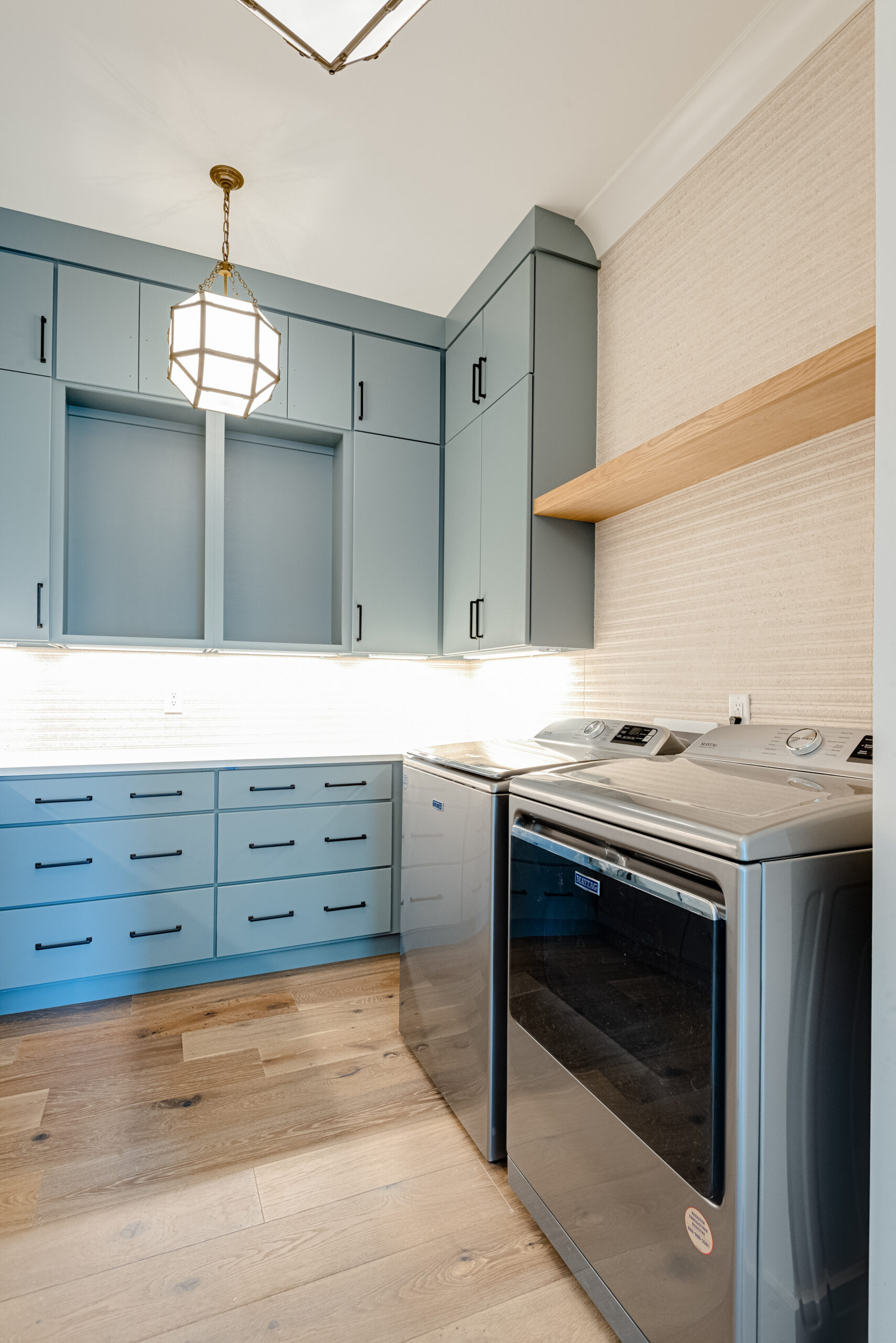 Laundry room with dusty blue cabinetry, tiled backsplash with wood stained open shelving