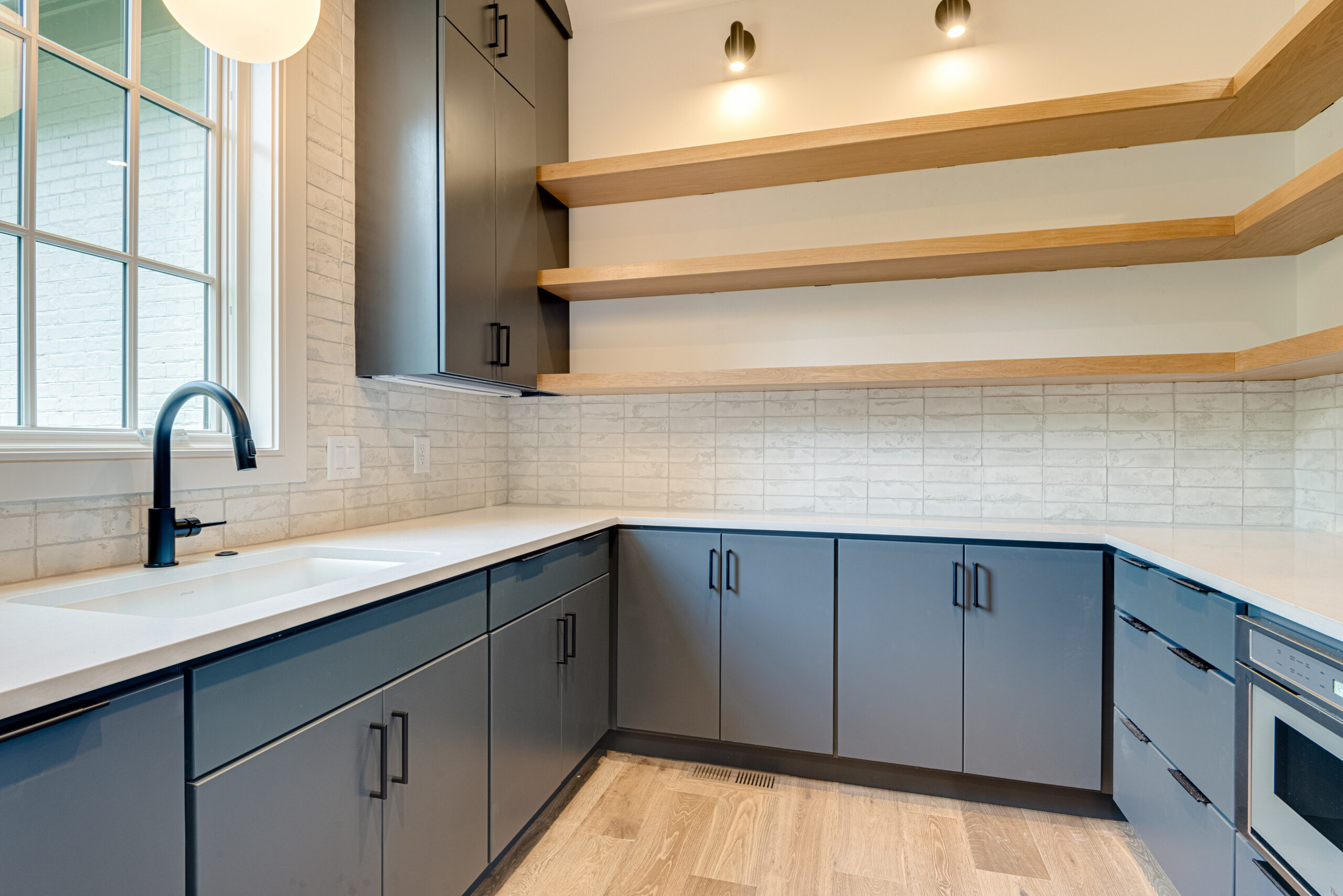 Navy cabinetry with light countertops and backspash, with light wood stained open shelving and sconce lighting