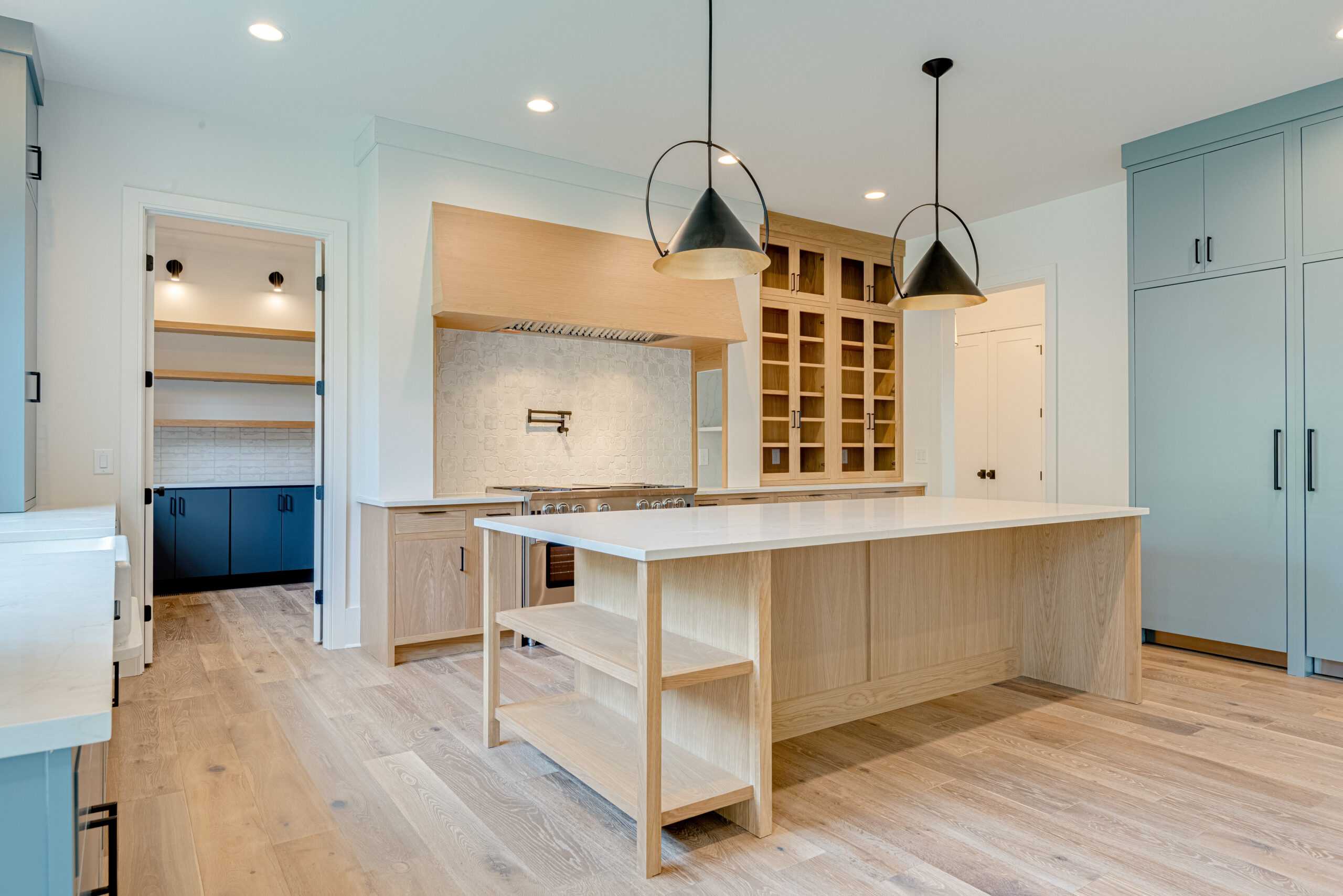 Kitchen with light blue cabinets, black dome pendants, white oak island, farmhouse sink, and glass-front pantry cabinets
