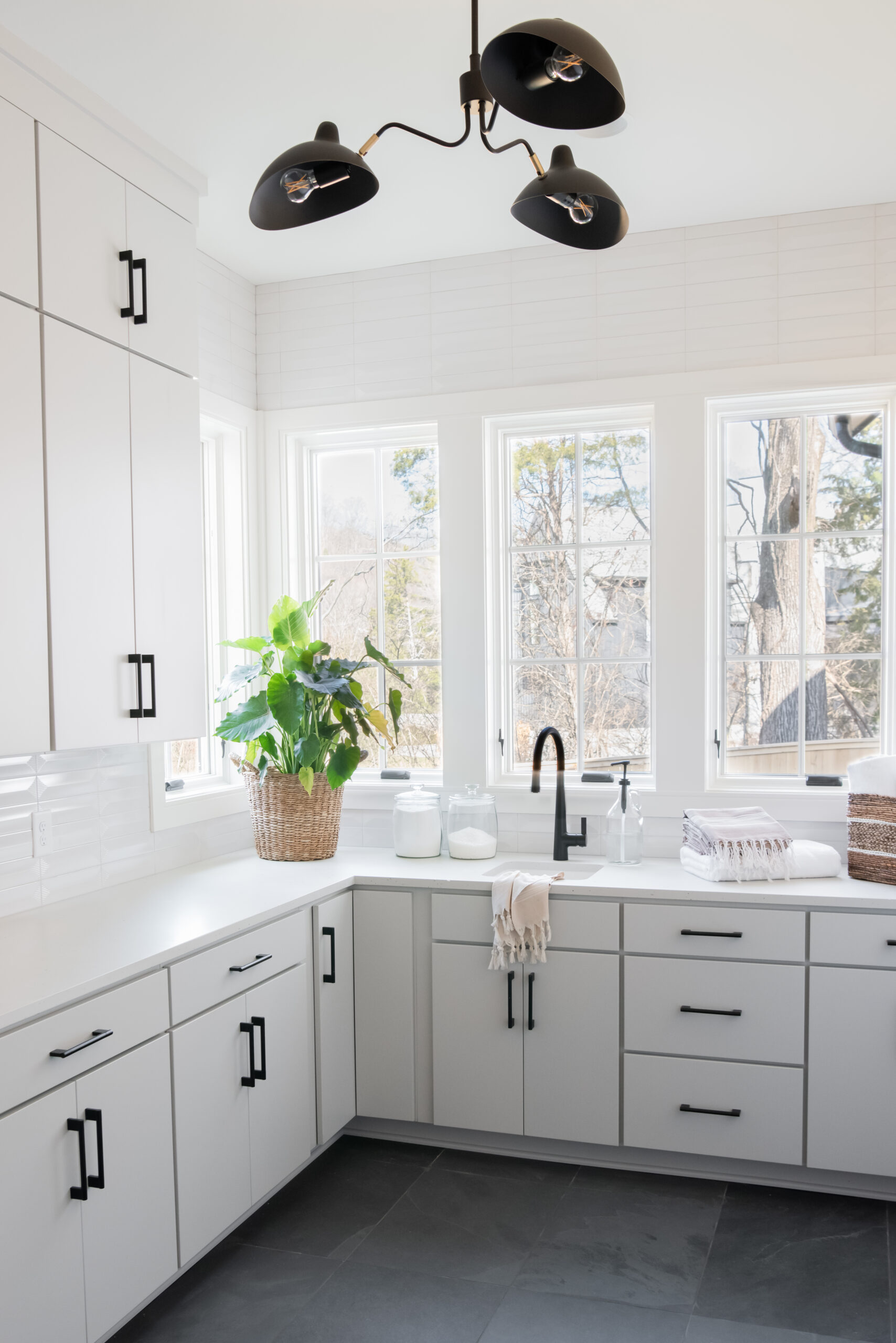 A large white laundry room with windows and white cabinetry below and black modern chandlier with black tiled flooring
