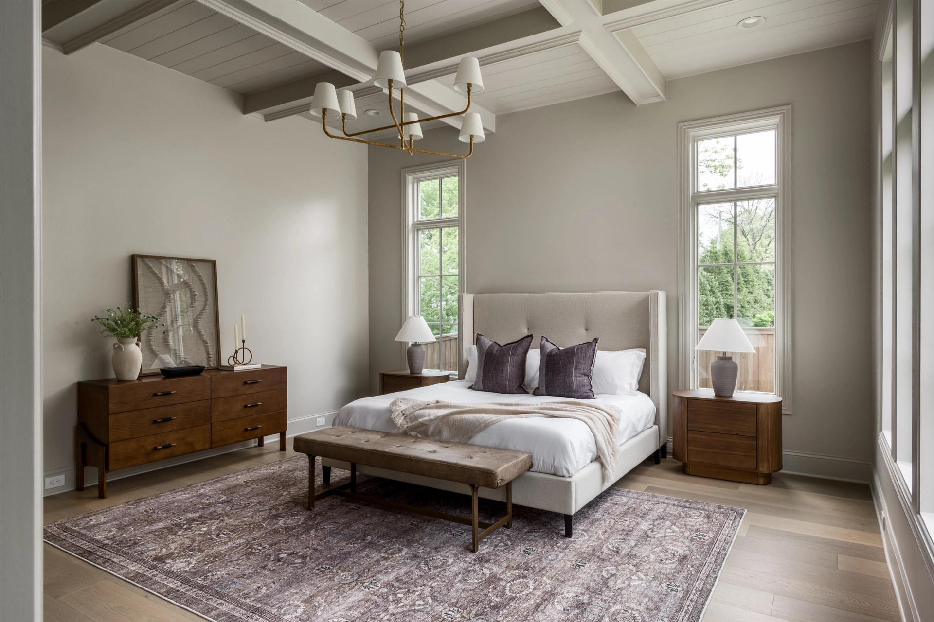 Bedroom with beamed ceiling, upholstered bed, brass chandelier, wood dresser, and a bench