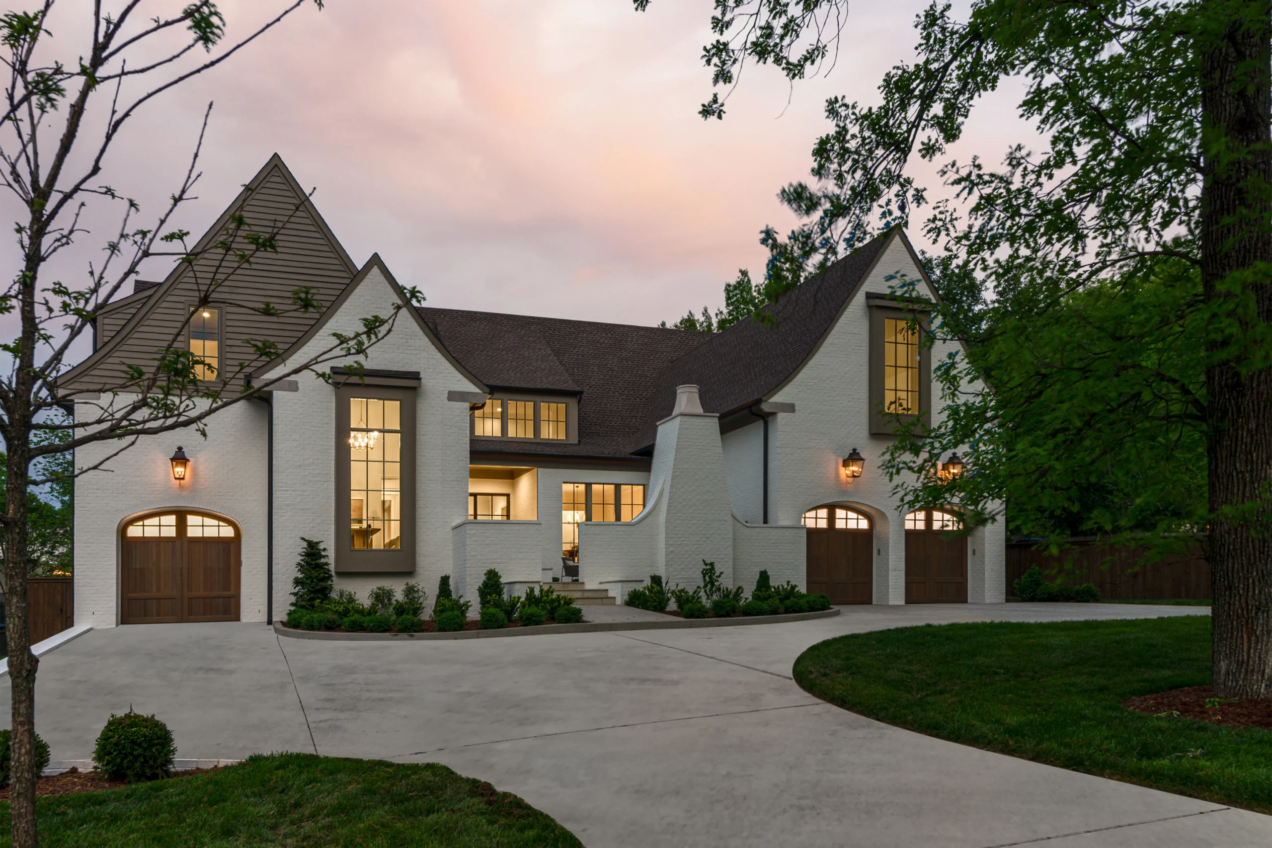 White brick Tudor-style home at dusk with arched garage doors and glowing windows under a sunset sky