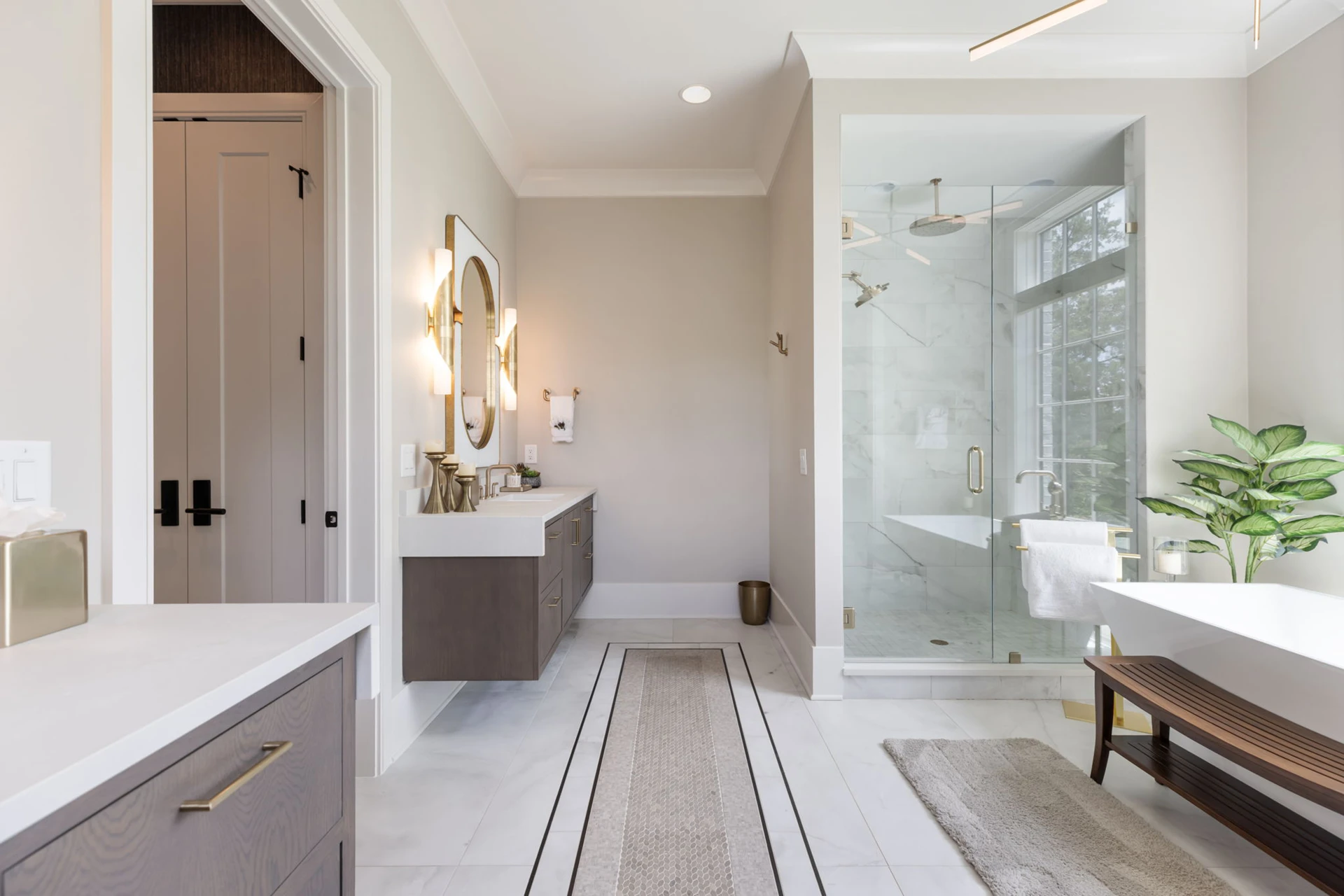 Master bath with gray floating vanity, gold oval mirror, marble glass shower, freestanding tub, and wood bench
