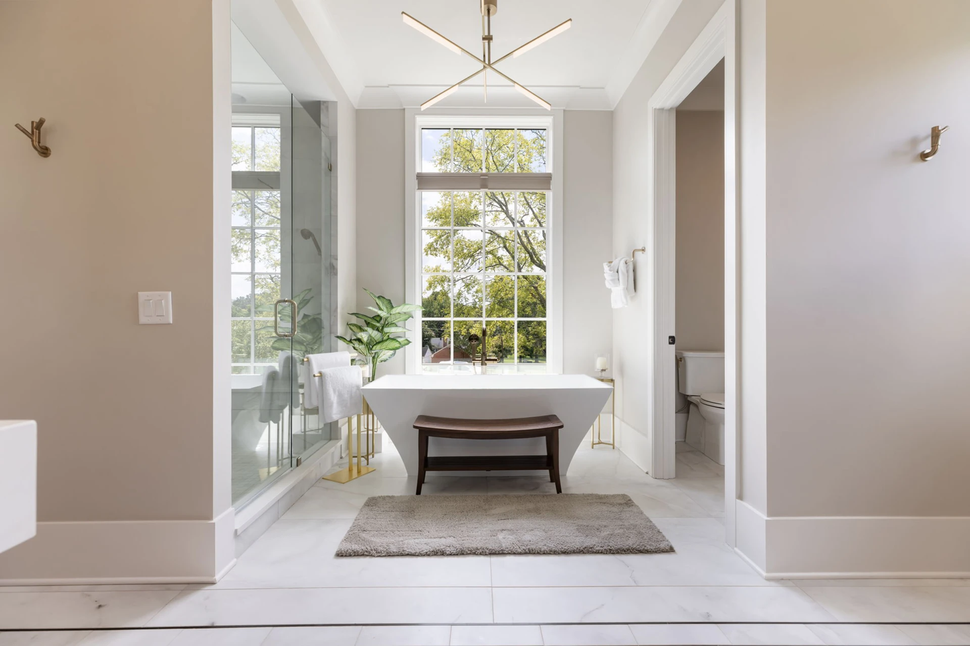 Master bath with freestanding tub, gold linear chandelier, glass shower, water closet, and a wood bench