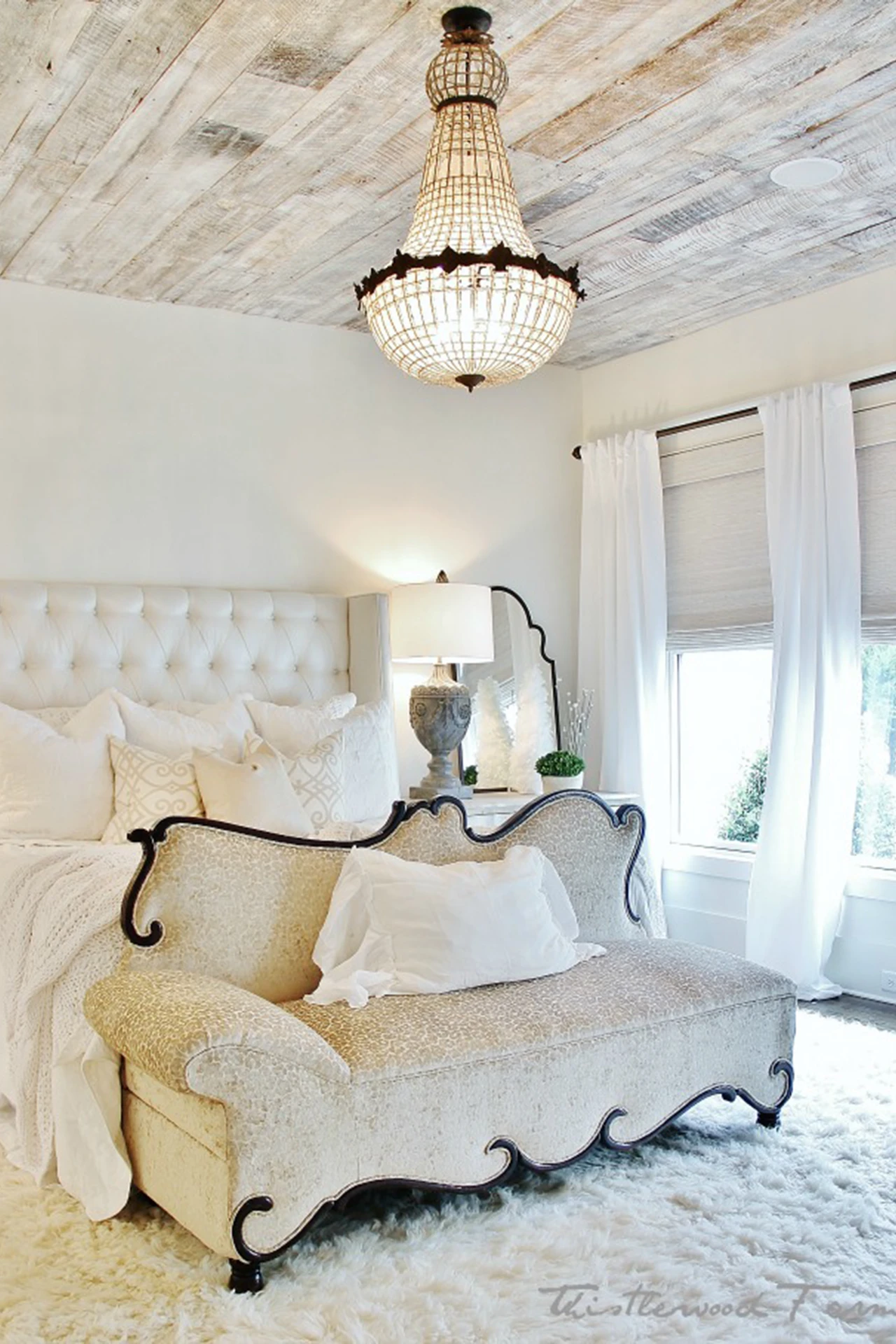White bedroom with whitewashed wood ceiling, iron bed with upholstered chaise, and a crystal chandelier
