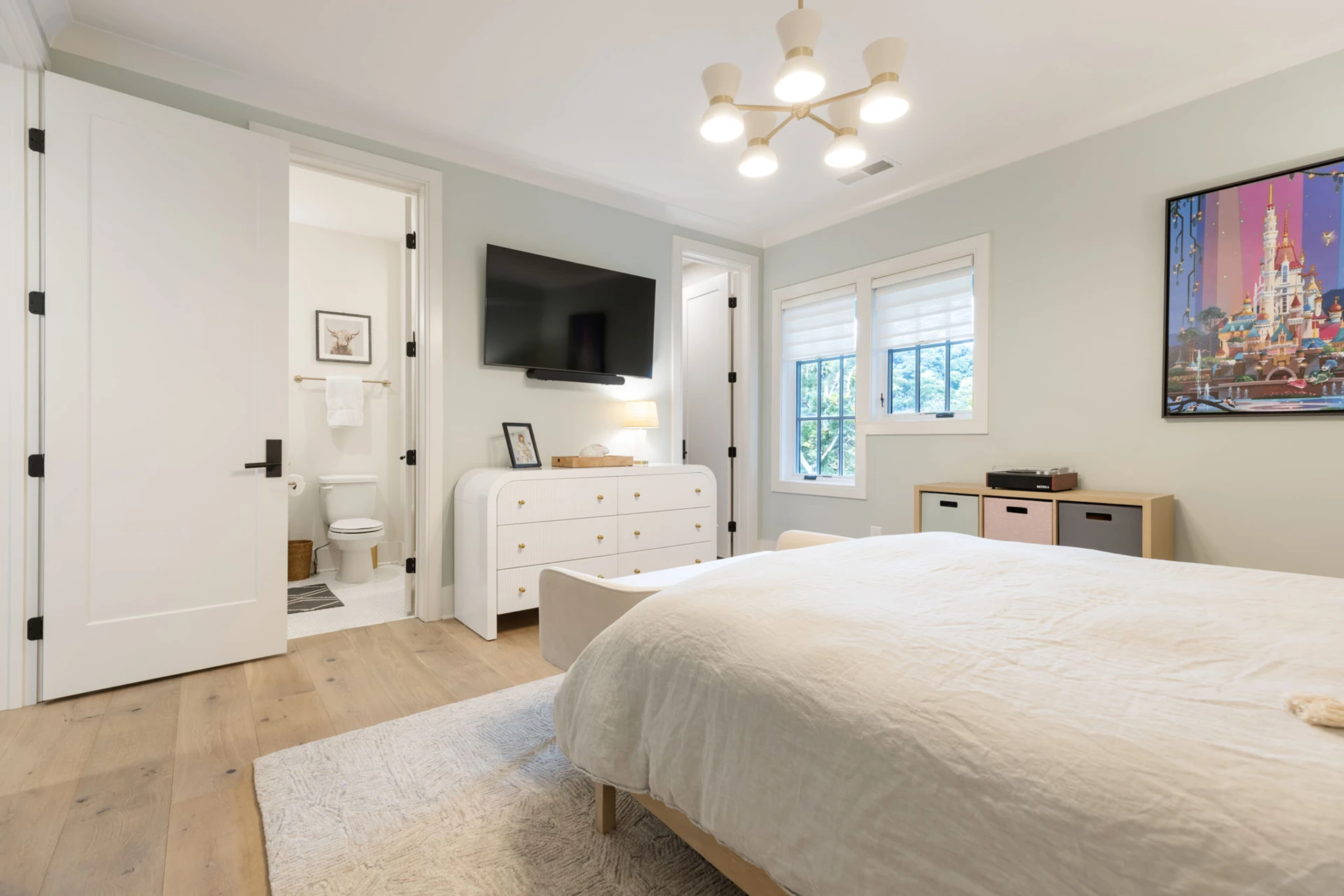 Bedroom with light green walls, wood bed, TV over a white dresser, and an ensuite bathroom