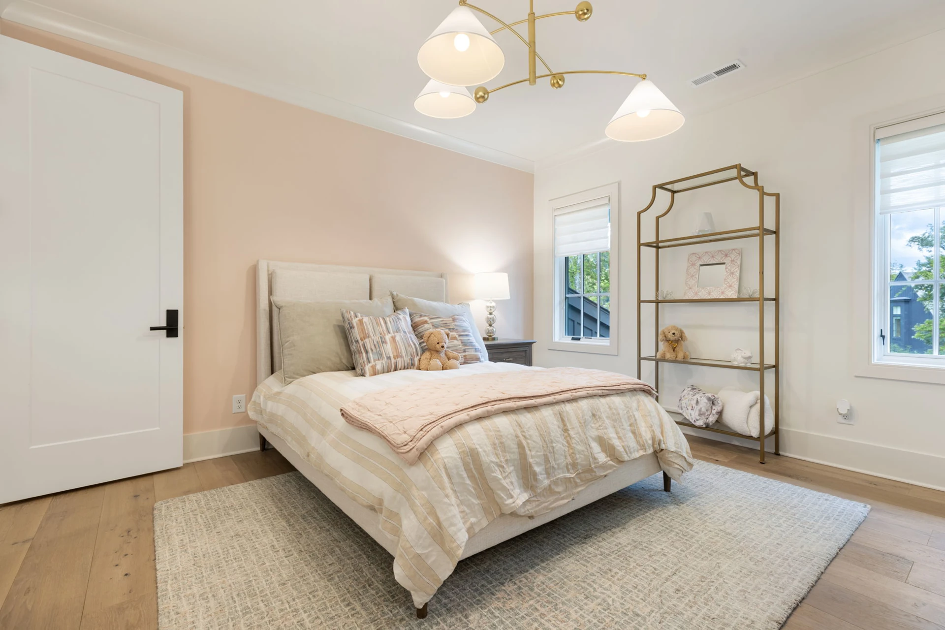 Bedroom with blush pink accent wall, upholstered bed, gold pendant light, and a gold étagère