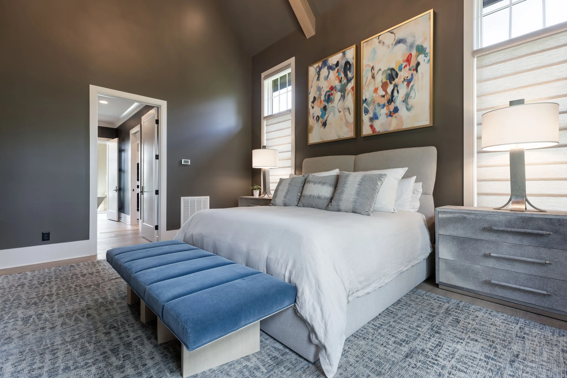 Bedroom with vaulted ceiling, taupe walls, upholstered bed, abstract diptych art, and a blue bench