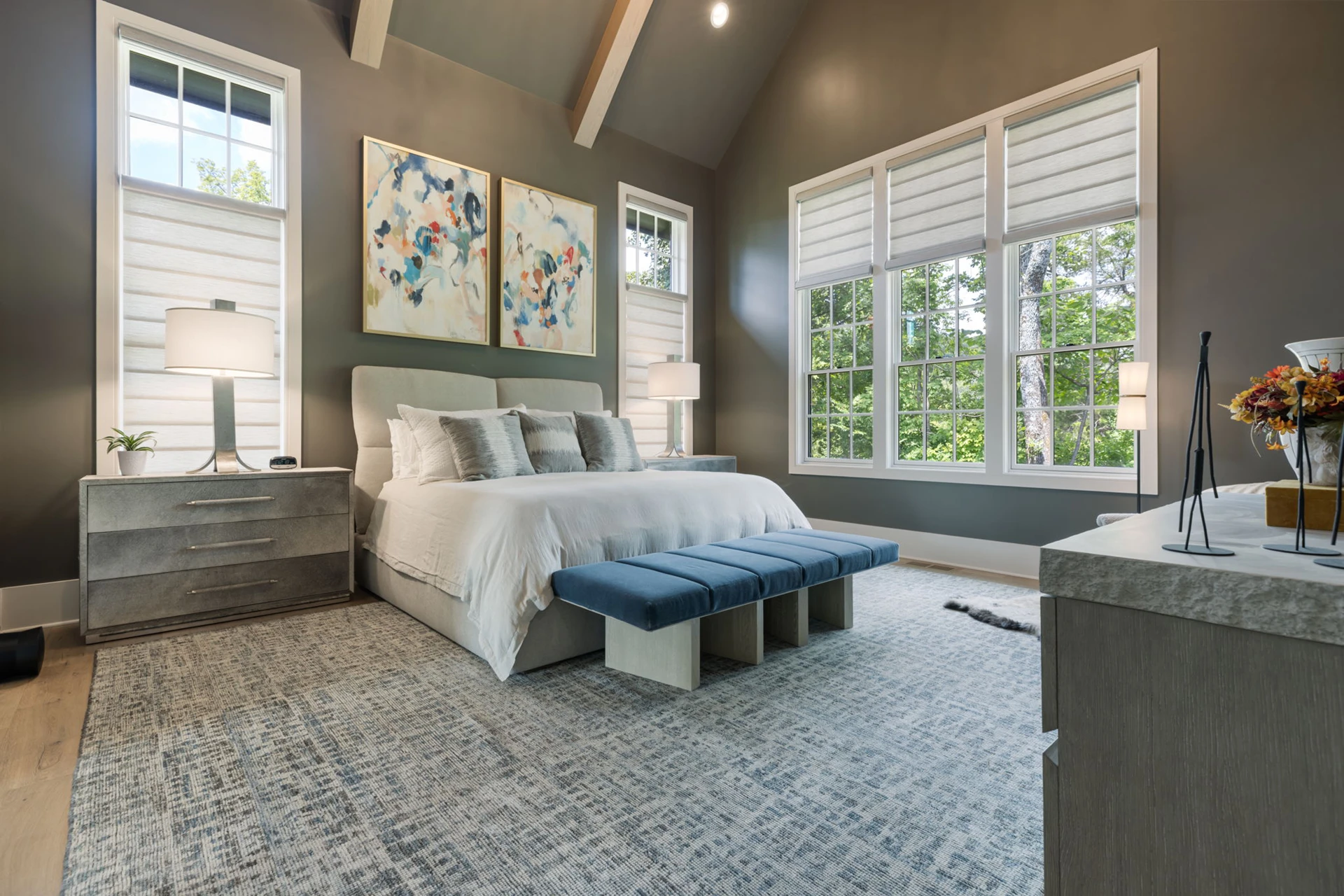 Bedroom with gray walls, vaulted beamed ceiling, upholstered bed, abstract art, and blue benches