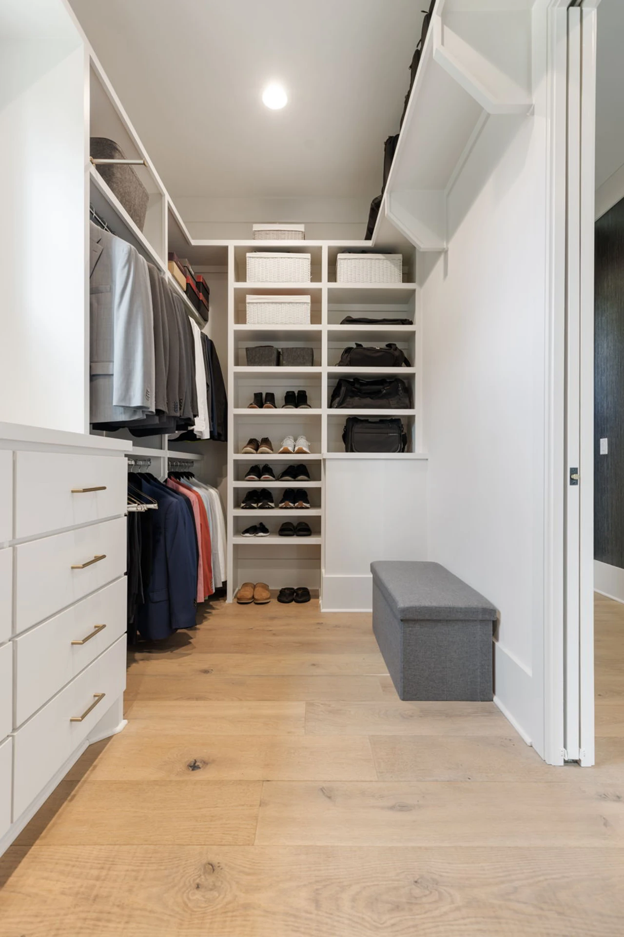 White walk-in closet with hanging rods, shelving, cubbies, and a gray upholstered storage bench