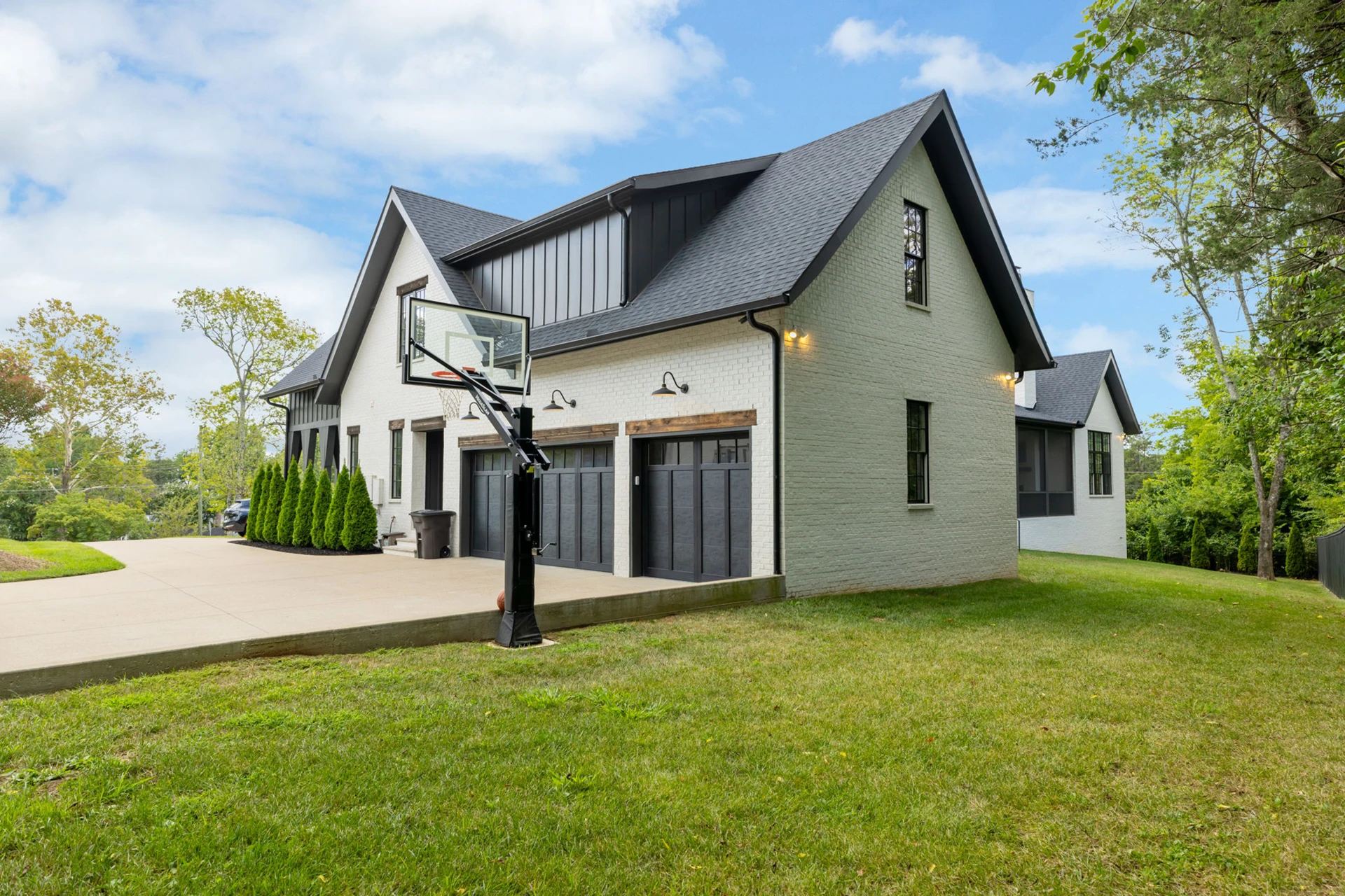 Side view of a white brick home with detached garage, dark garage doors, and a driveway basketball hoop