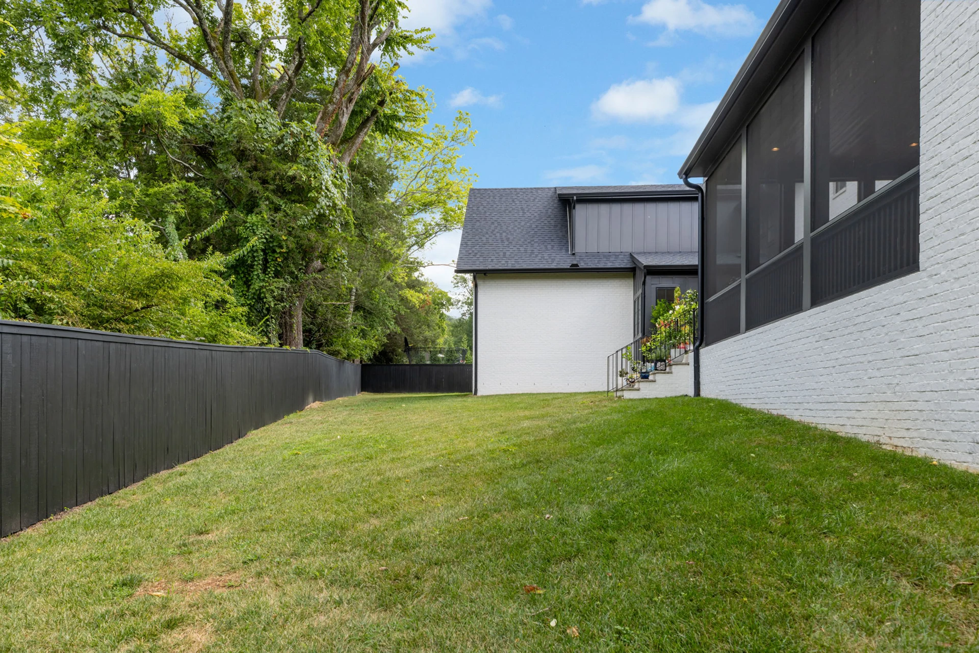 Side yard of a white brick home with a black privacy fence, green lawn, and screened porch