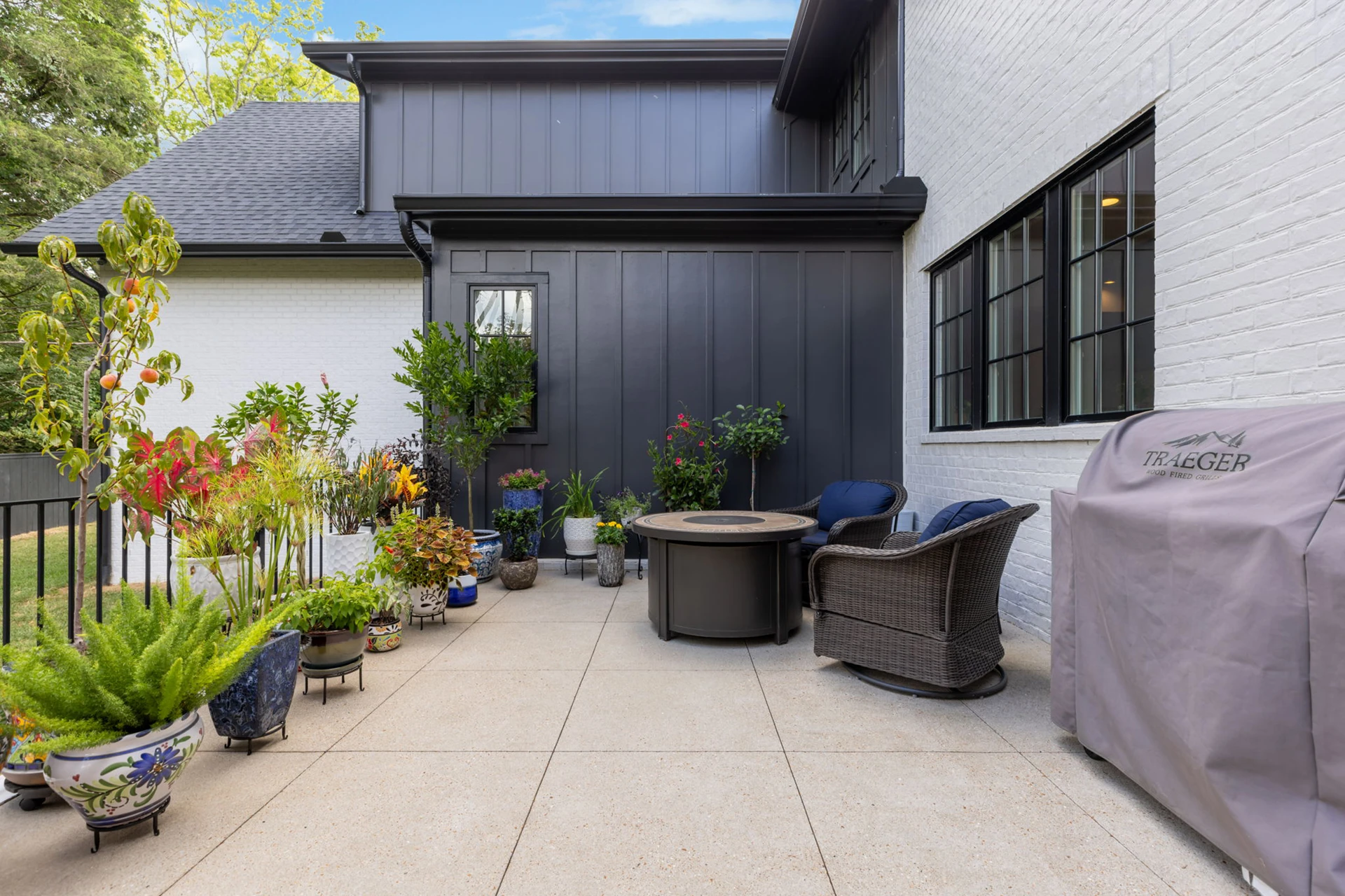 Courtyard patio with dark board-and-batten wall, potted plants, fire pit with wicker chairs, and a covered grill