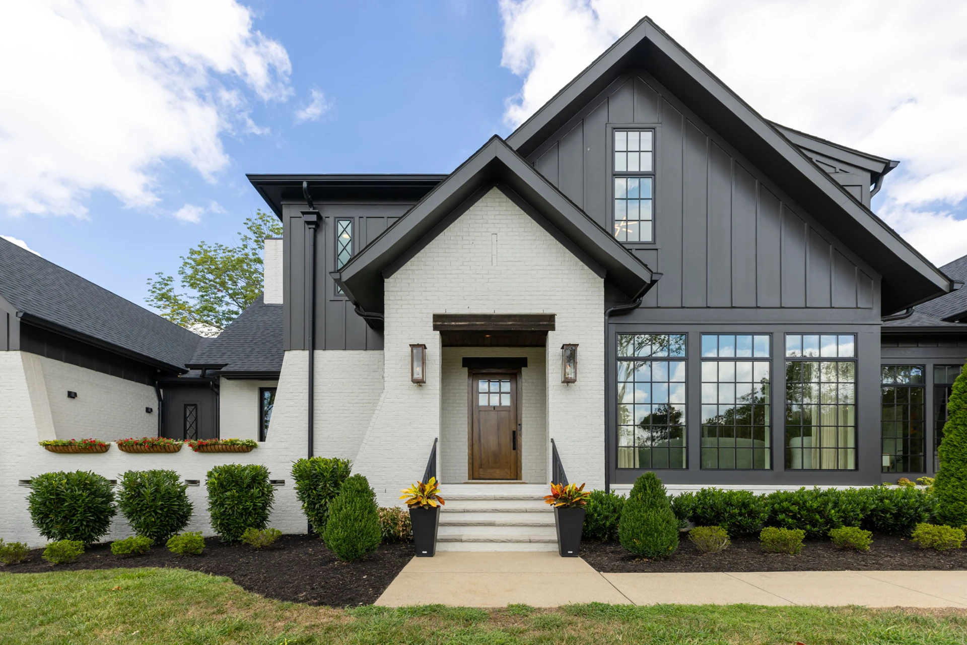 White brick and dark board-and-batten home entry with a gabled porch, wood door, and landscaped beds
