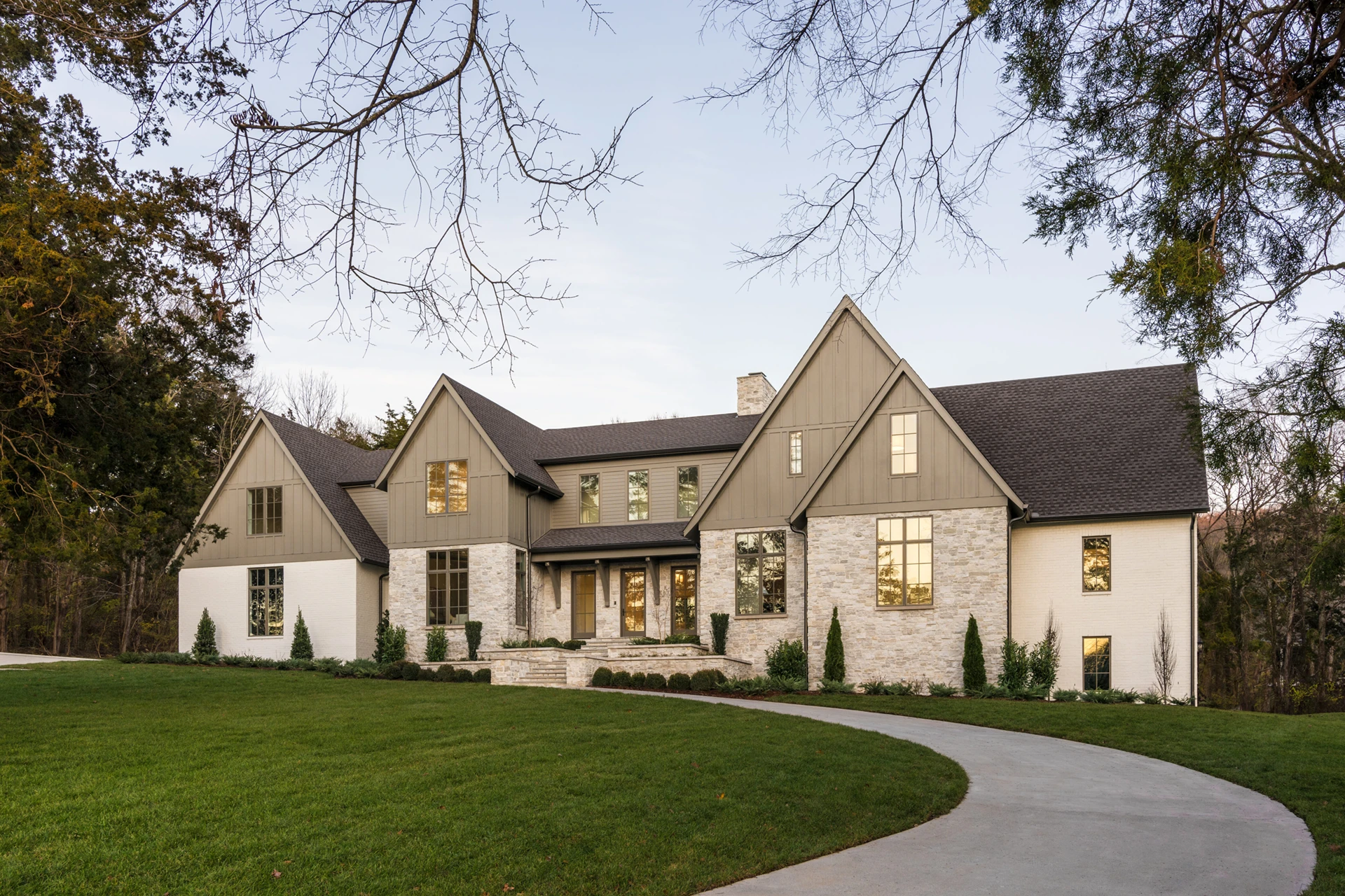 Two-story custom home with stone and board-and-batten siding lit at dusk, with a curved driveway