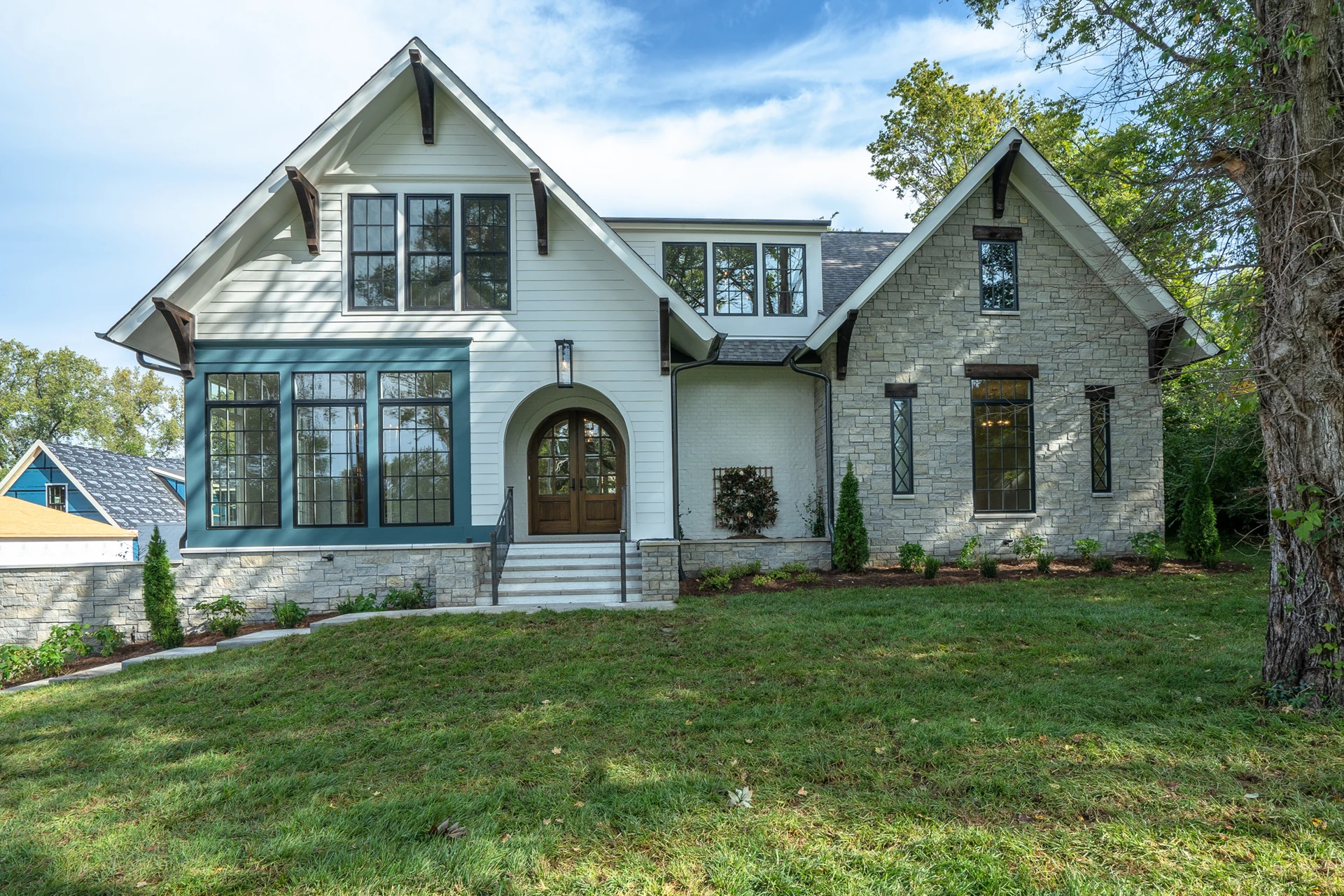 White and gray home with stone accents, arched wood entry, and teal-framed bay windows