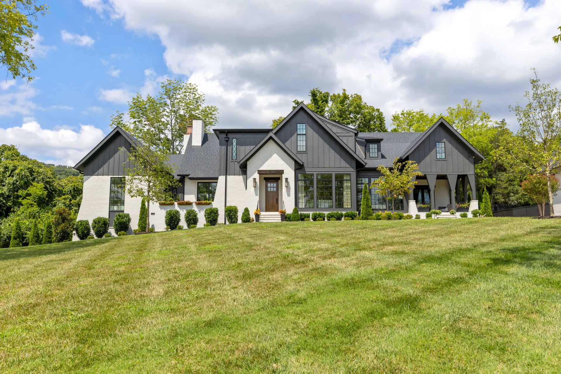 White brick and dark board-and-batten home on a hill with dark roof, multiple gables, and a wide lawn