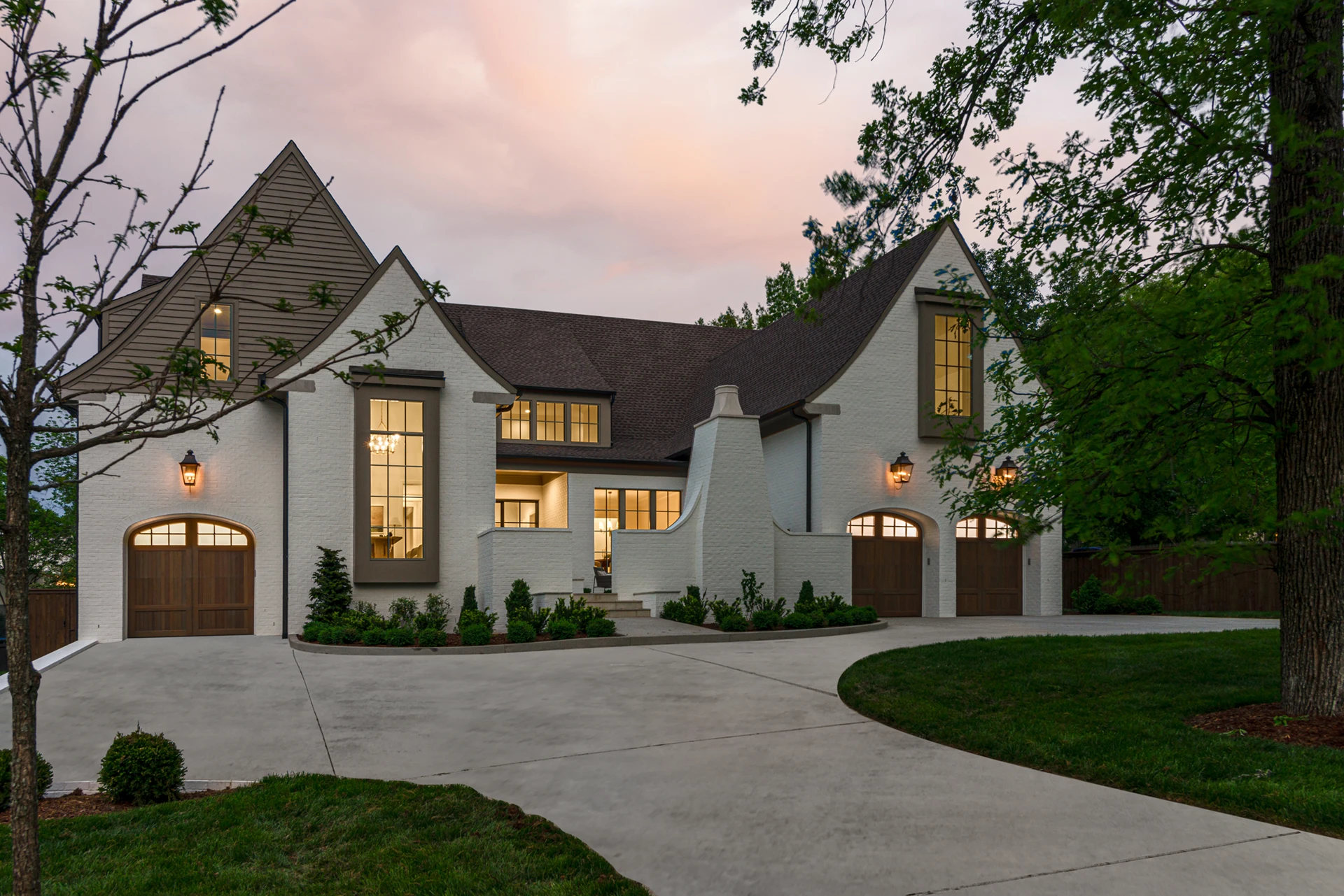 White brick Tudor-style home at dusk with arched wood garage doors and glowing windows under a sunset sky