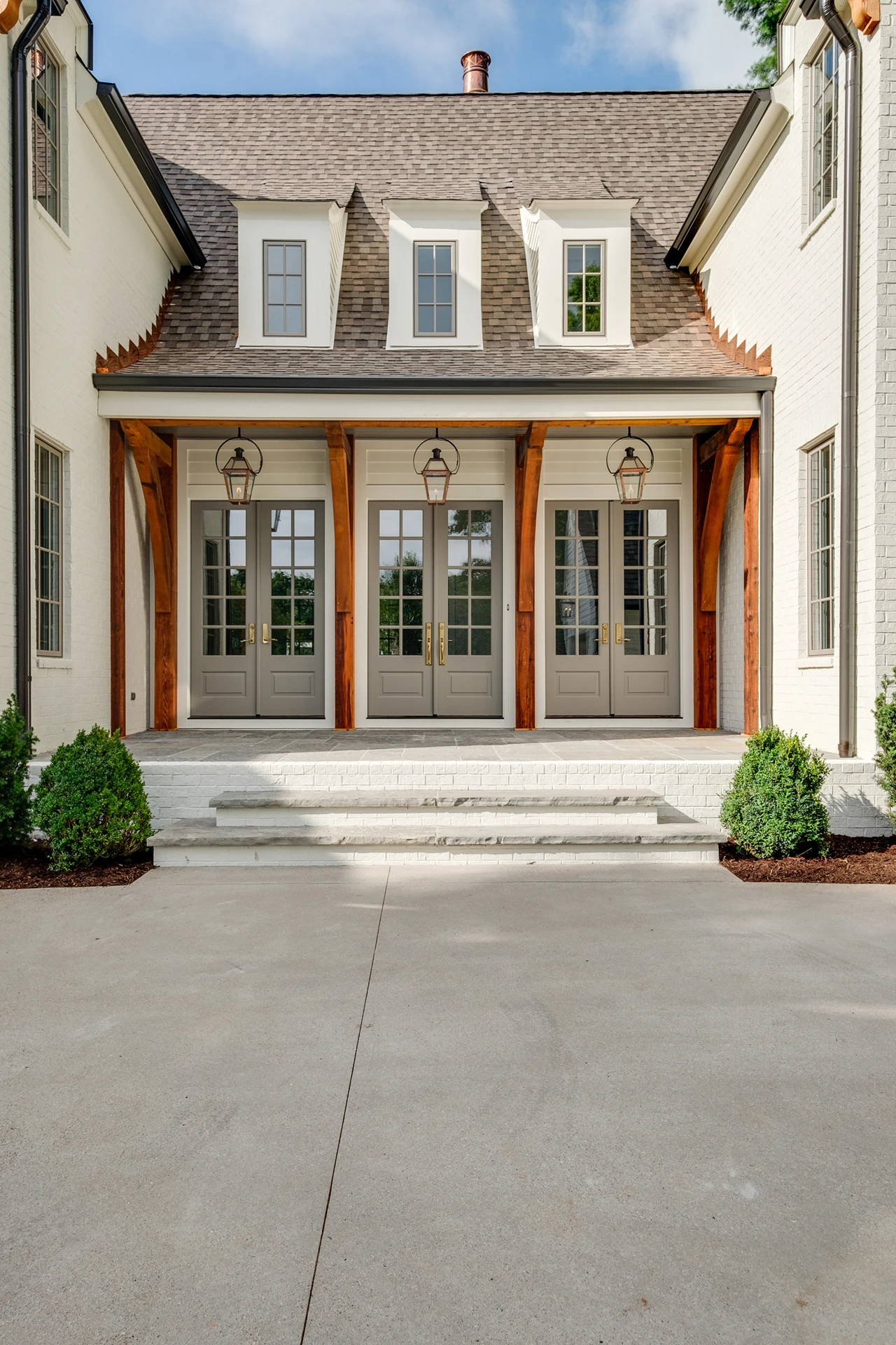 White home entry with dormer windows, wood columns, three wood-and-glass doors, and a paver driveway