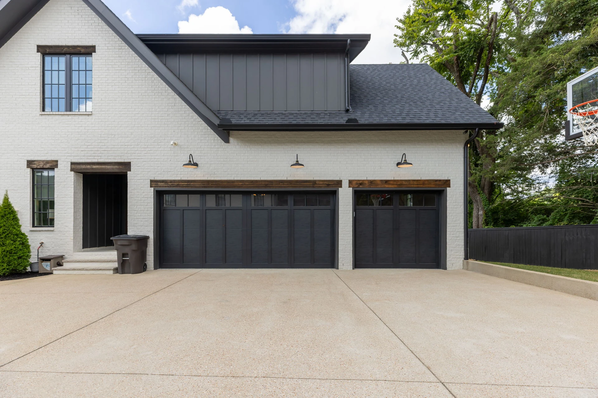 White brick home with black board-and-batten upper, two black garage doors with wood headers, and a driveway