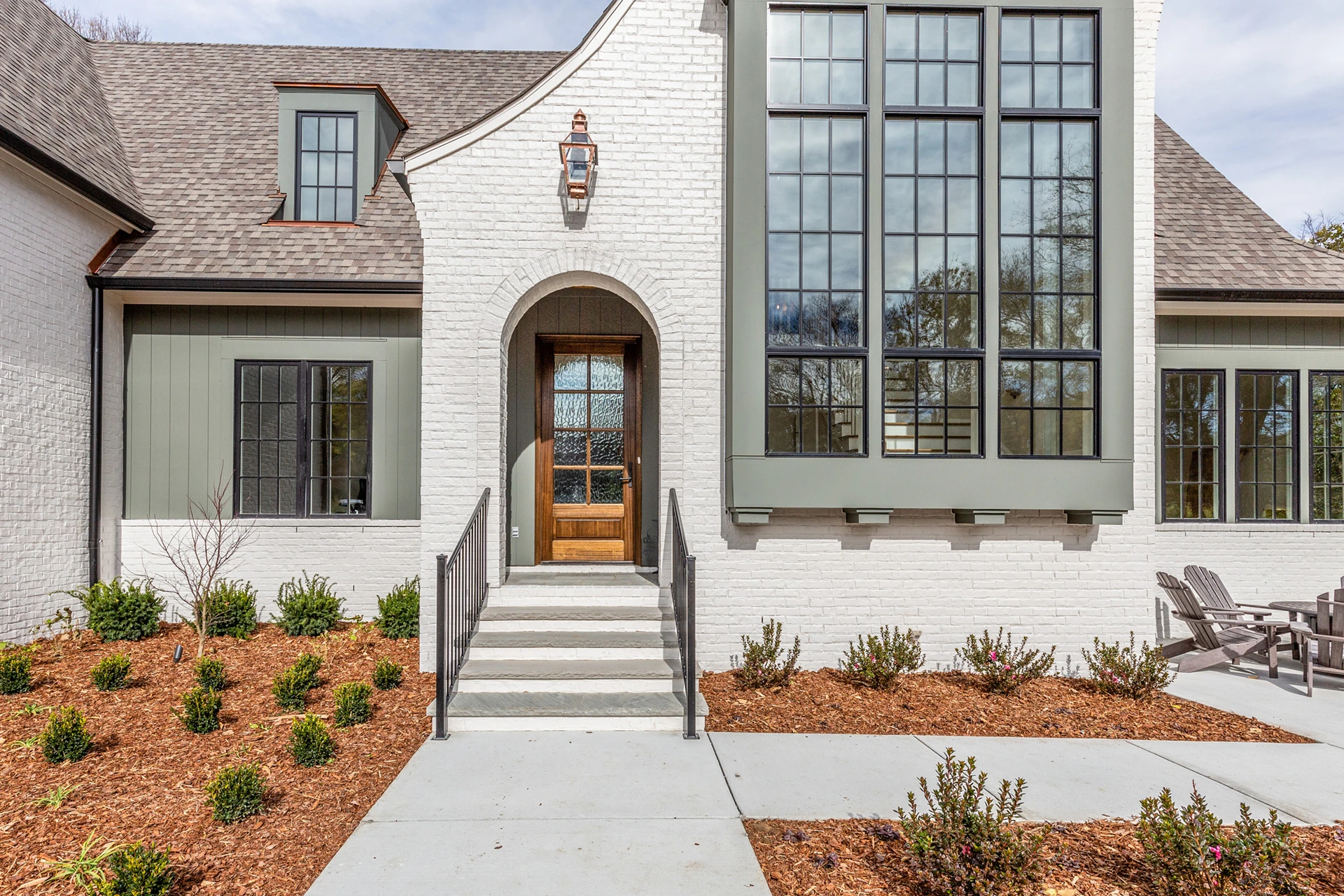 White brick home entry with arched wood door, sage accents, tall black-grid windows, and Adirondack chairs