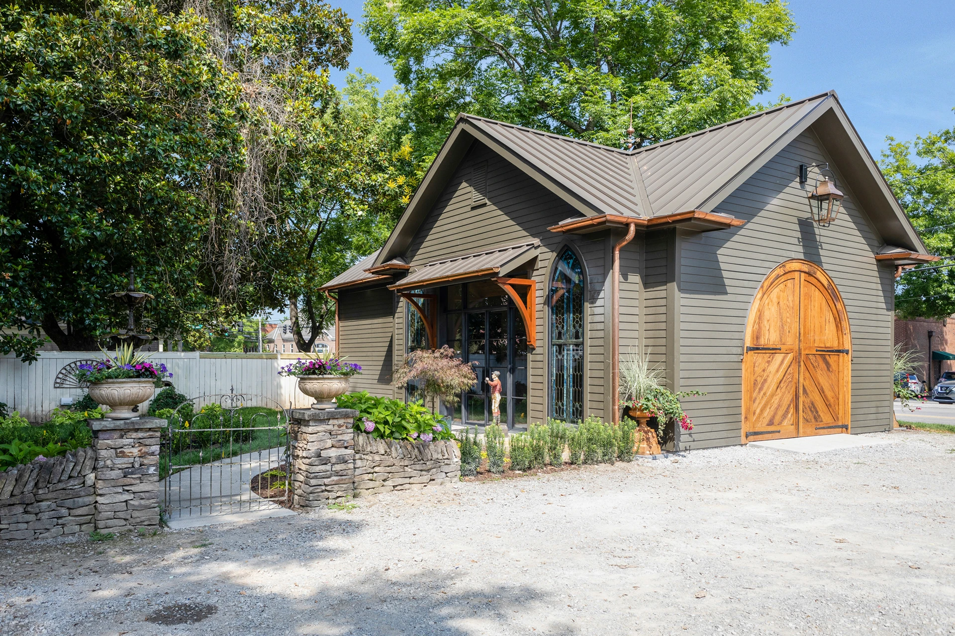 Tan cottage-style building with metal roof, arched wood doors, arched windows, and flower planters