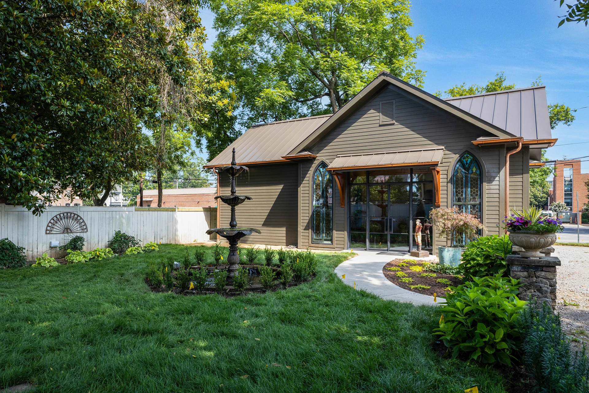 Tan cottage-style building with metal roof and arched windows, with a tiered fountain in a landscaped yard