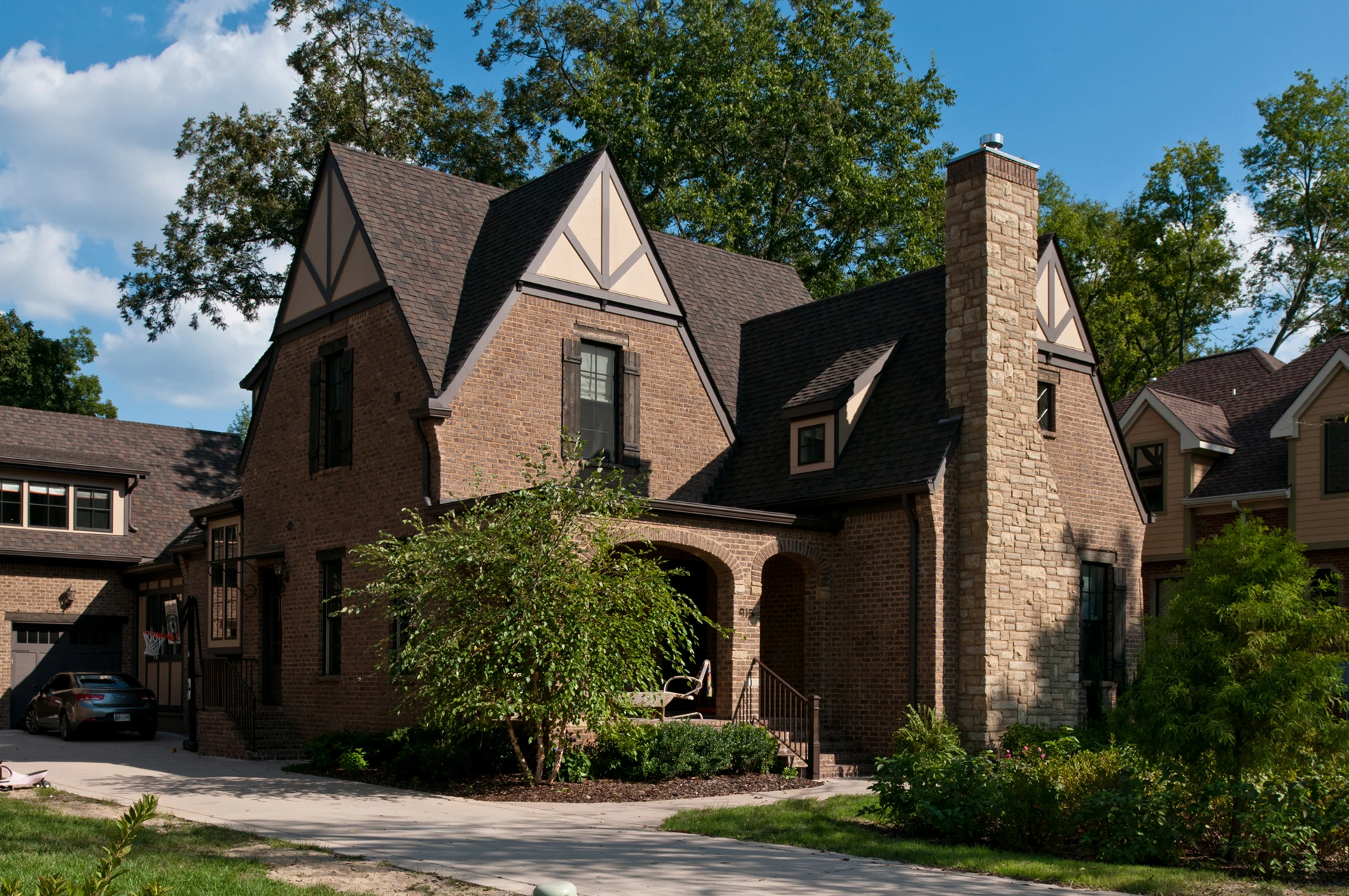 Brick Tudor-style two-story home with steep gable, stone chimney, arched entry, and mature landscaping