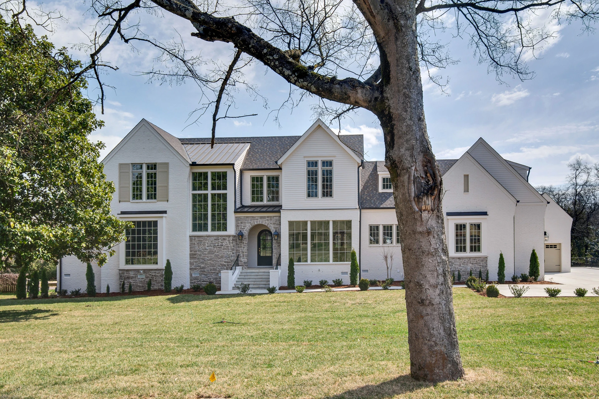 White two-story home with stone accents and arched entry, framed by a large bare tree in winter