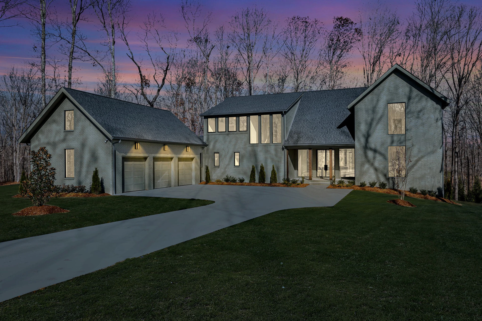 Gray stucco two-story home at dusk with attached garage, glowing windows, under a purple sunset sky