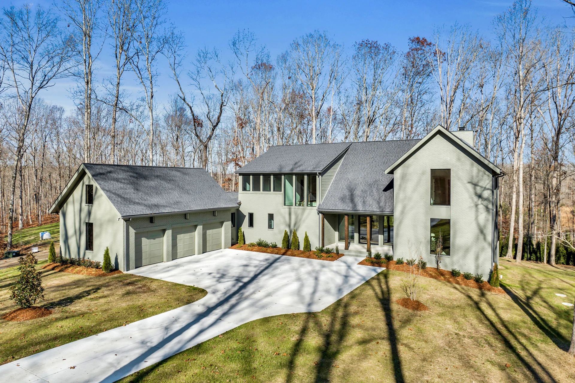 Aerial view of a gray modern home with attached three-car garage and a long driveway on a wooded lot
