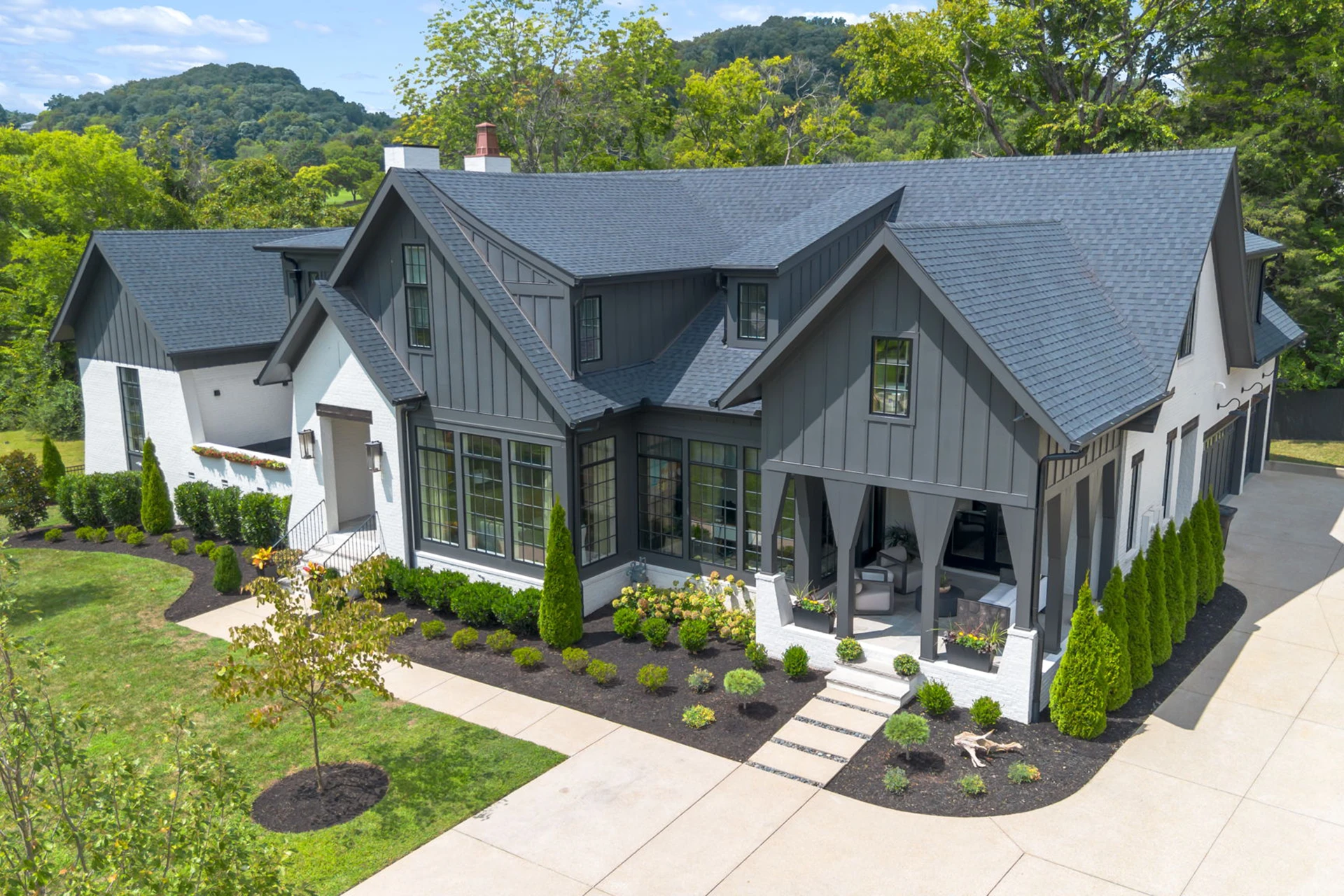 Elevated view of a gray and white modern farmhouse with dark roof, landscaped yard, and hills behind