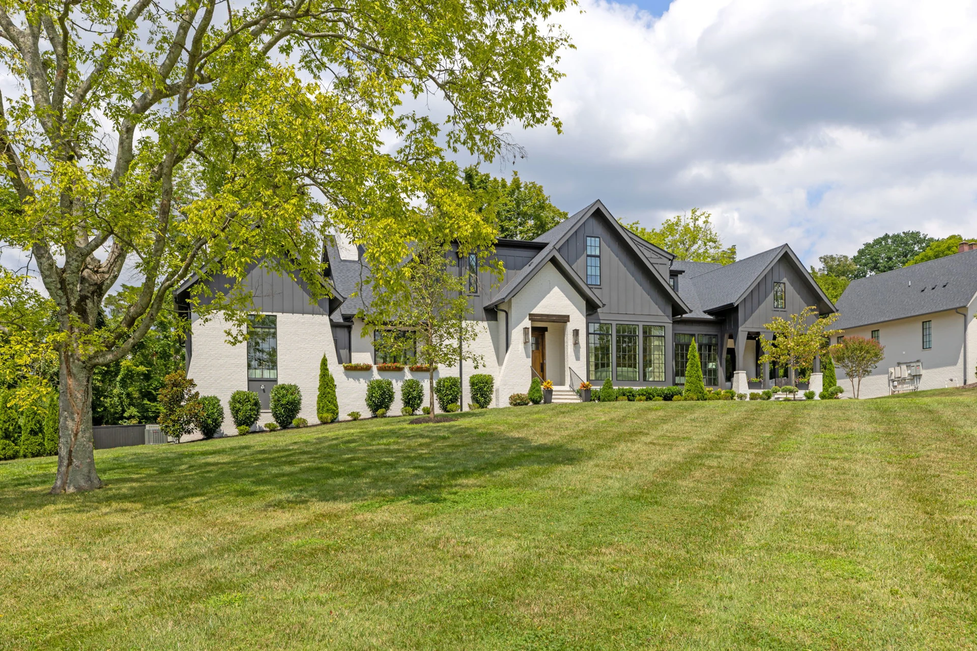 White brick and dark board-and-batten single-story home with a large shade tree on the front lawn