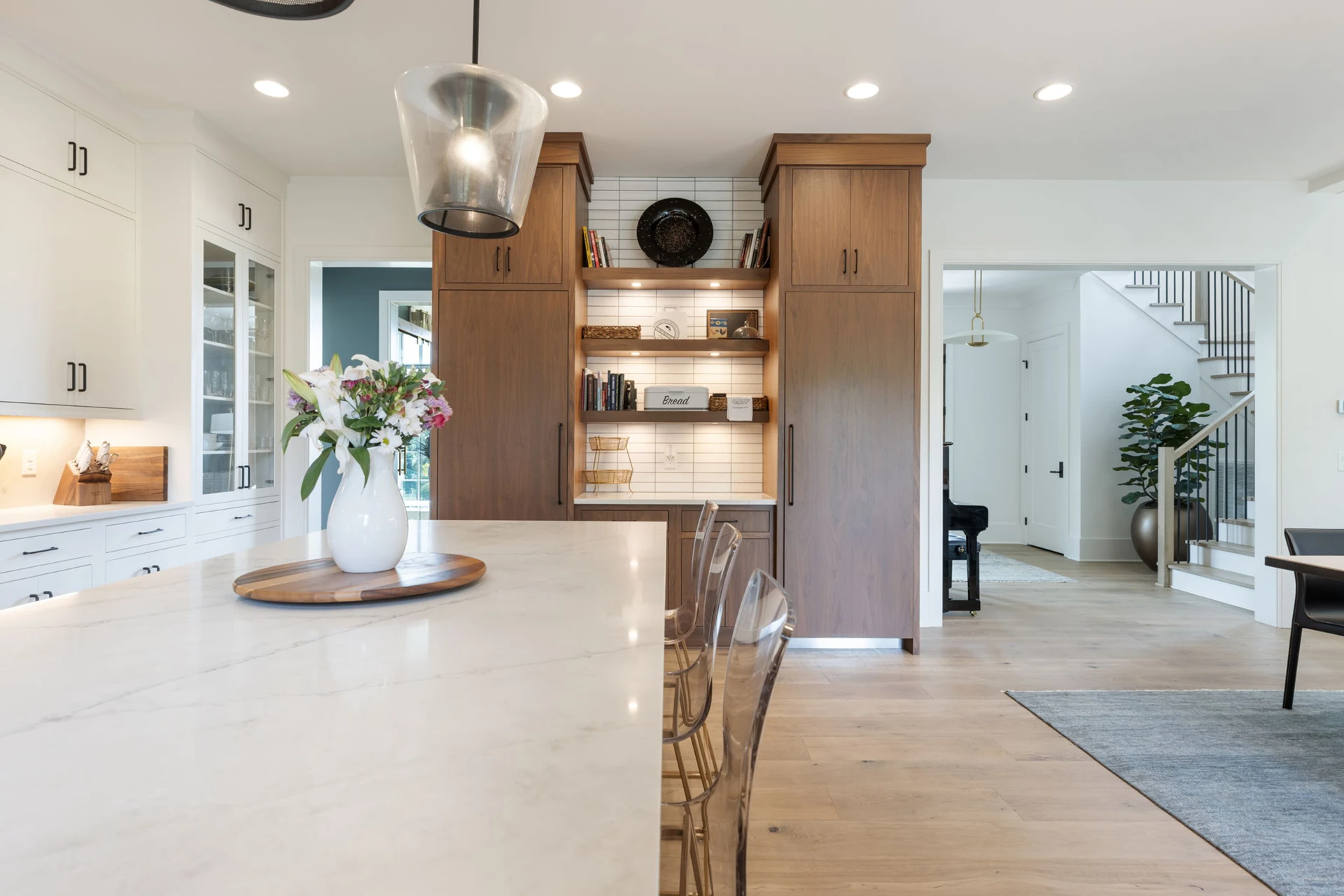 Kitchen island with marble waterfall counter, glass pendant, and vase of flowers, with wood pantry and staircase beyond