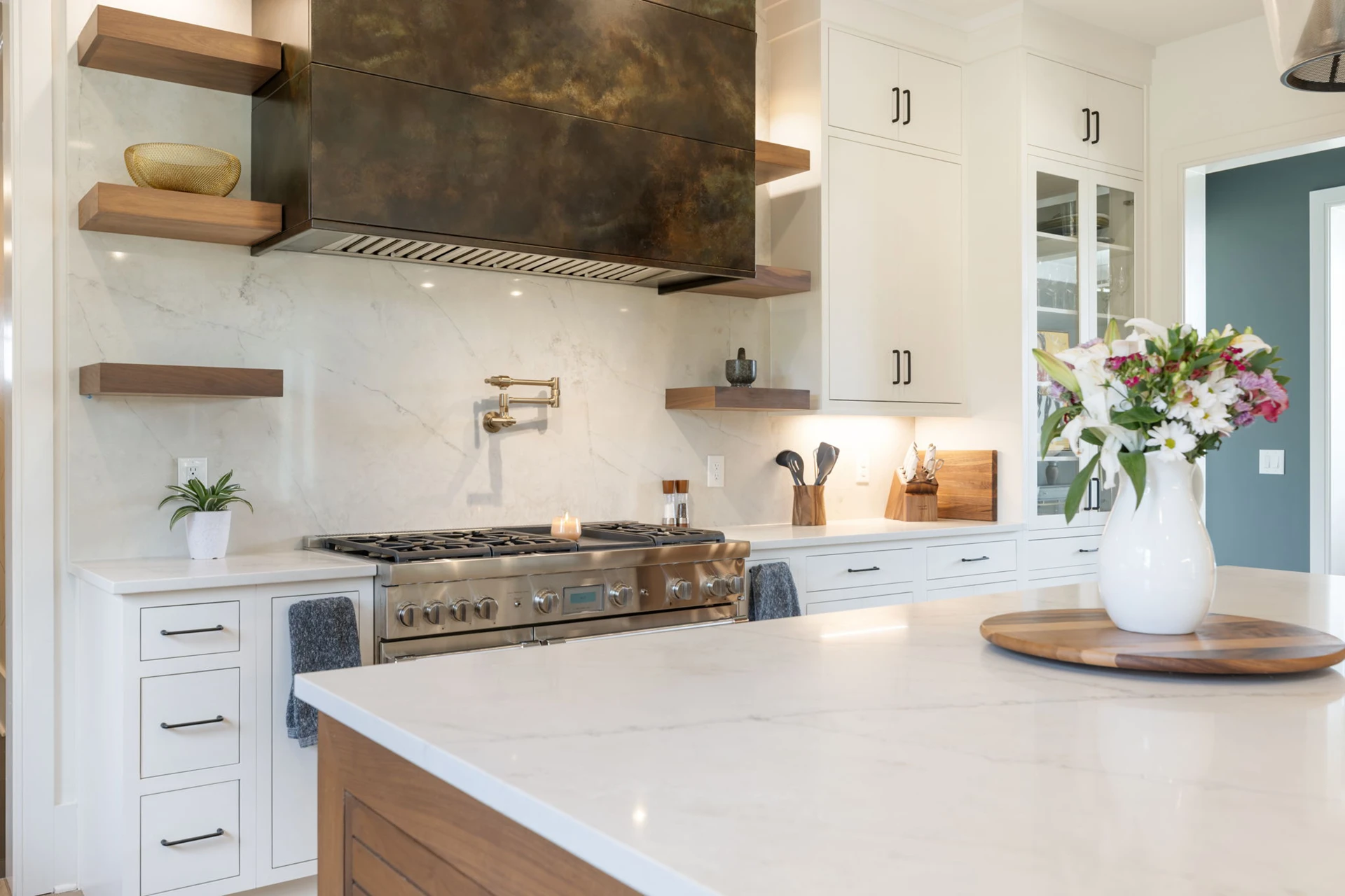 Kitchen with dark metal range hood, marble backsplash, floating wood shelves, and a marble island with wood base