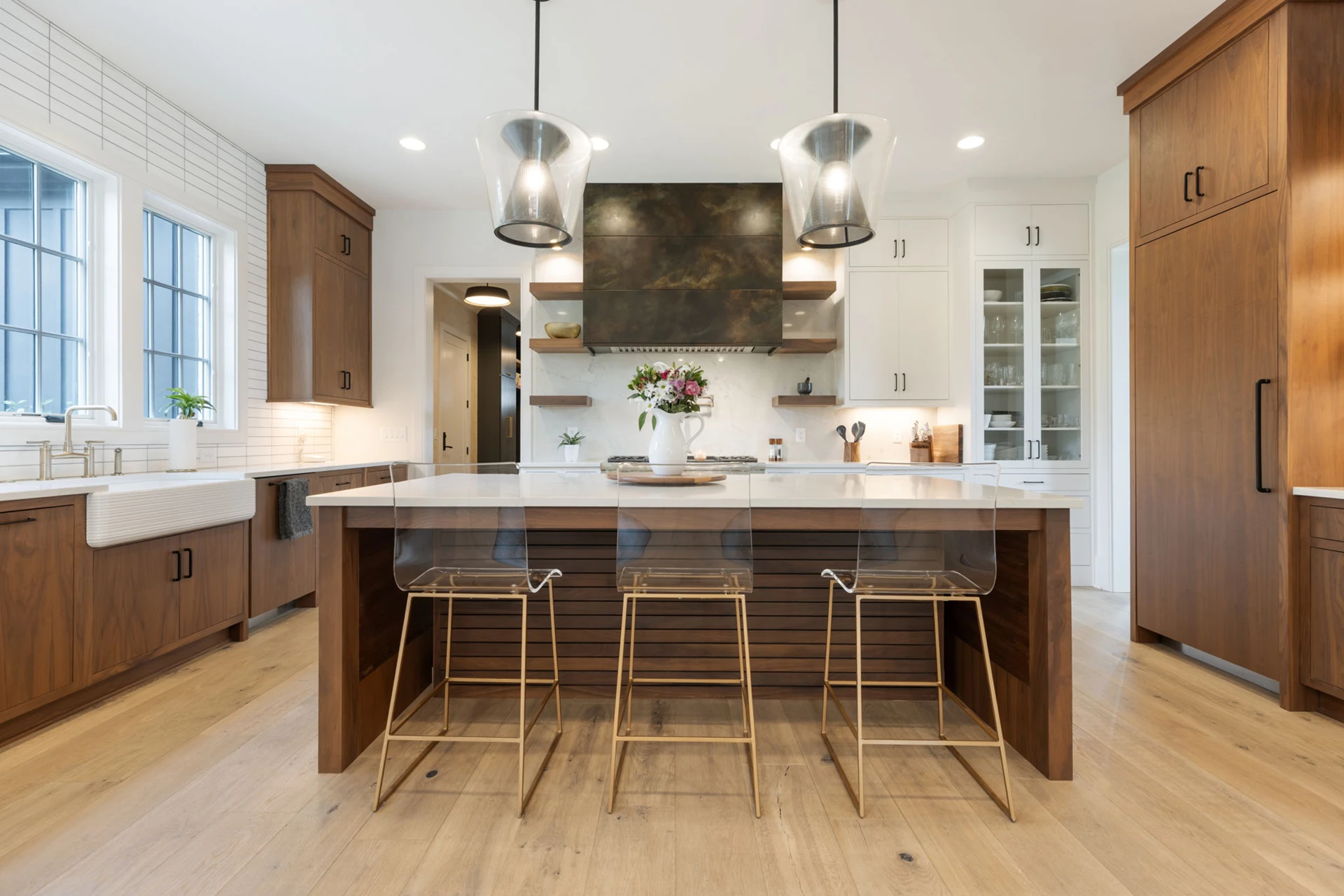 Kitchen with dark metallic range hood, wood and white cabinets, farmhouse sink, and a wood island with brass-legged stools