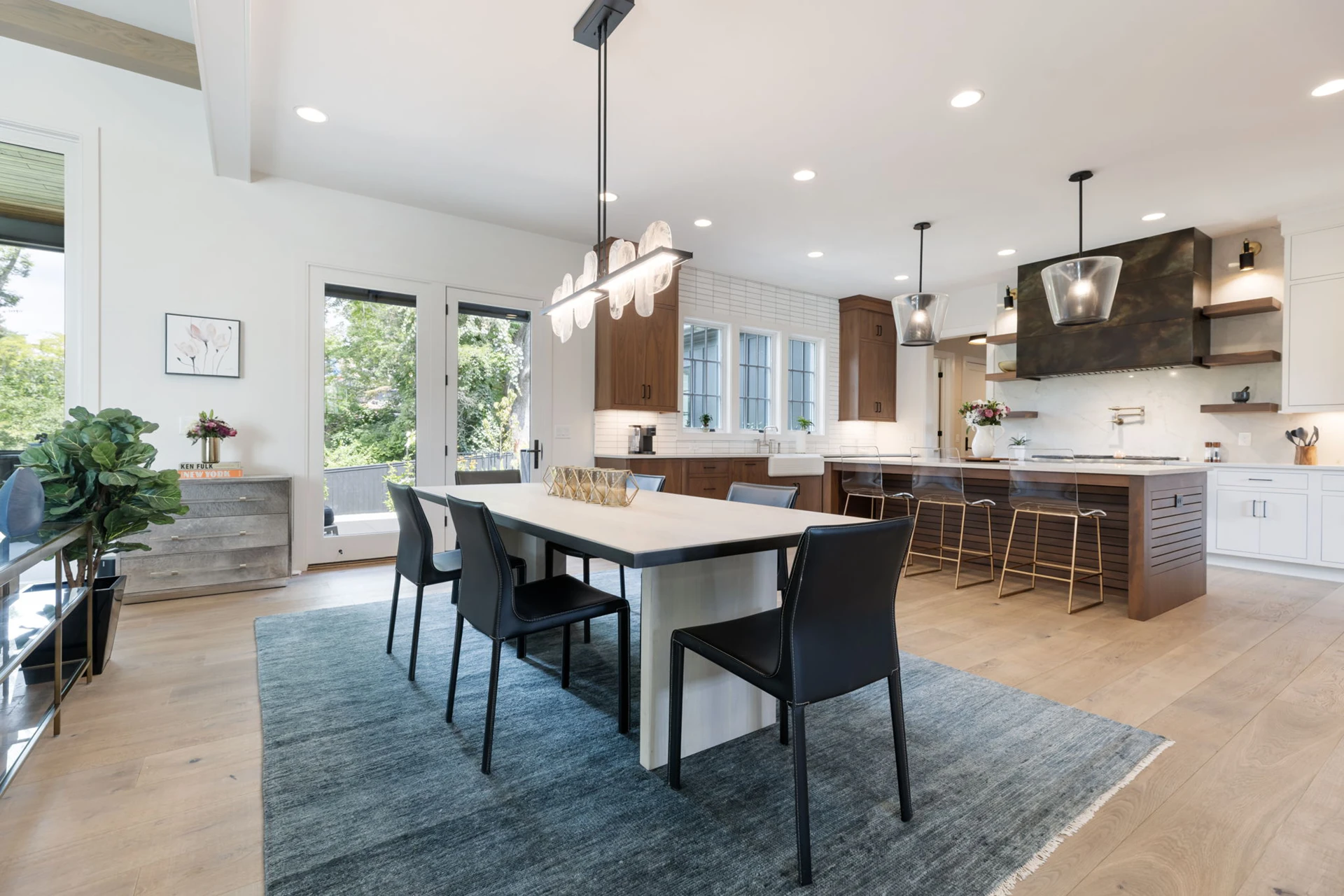 Open dining and kitchen with gray rug, white table with black chairs, linear chandelier, and a dark range hood beyond