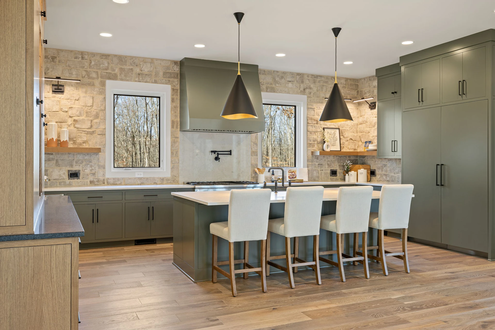 Kitchen with sage green cabinets, stone accent wall, black cone pendants, and cream upholstered island stools