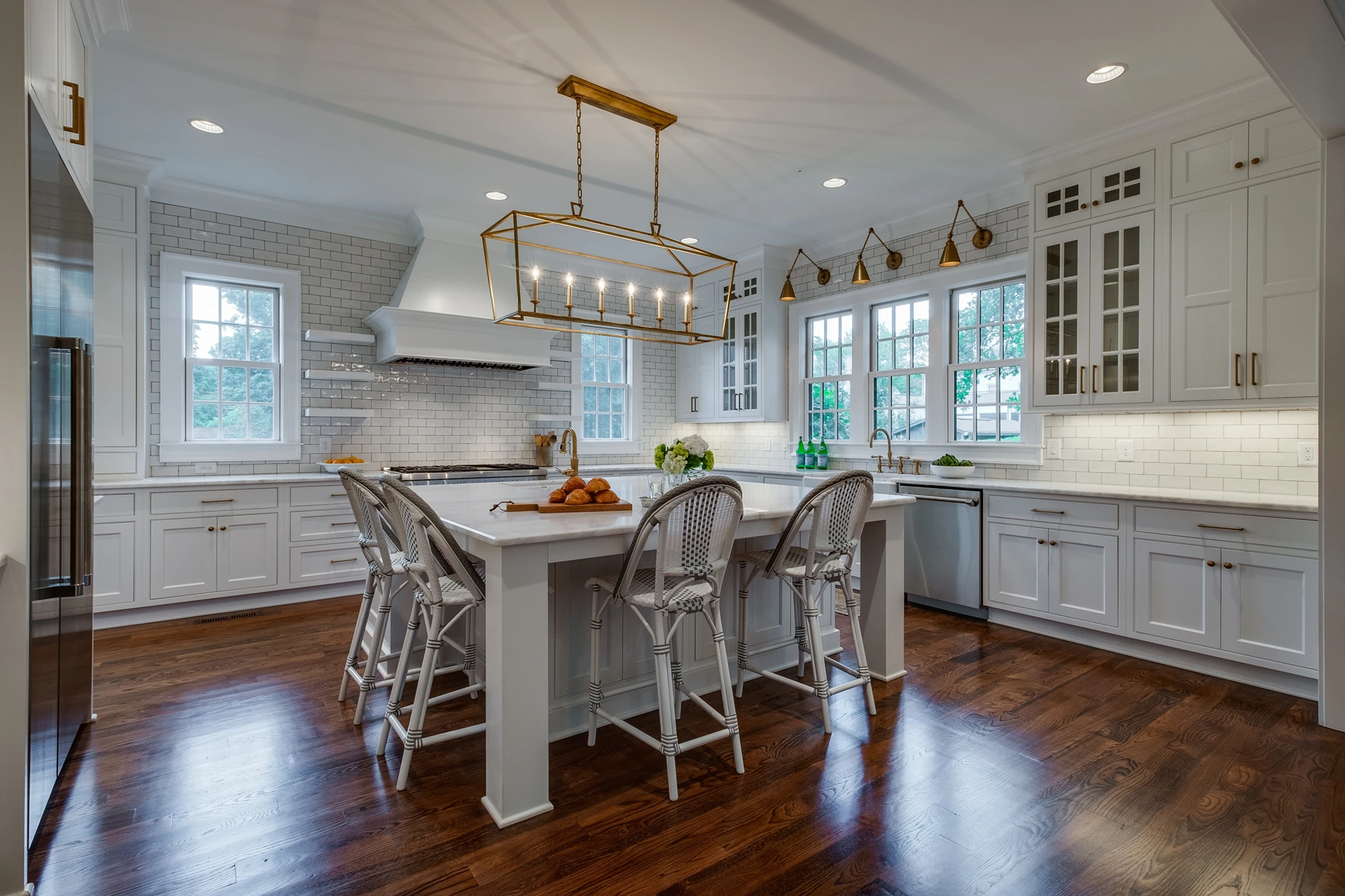White kitchen with brick backsplash, gold linear chandelier, woven counter stools, and dark wood floor