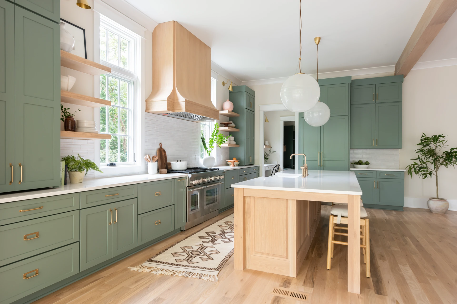 Green kitchen with wood range hood and island, open shelving, globe pendants, plants, and a patterned rug