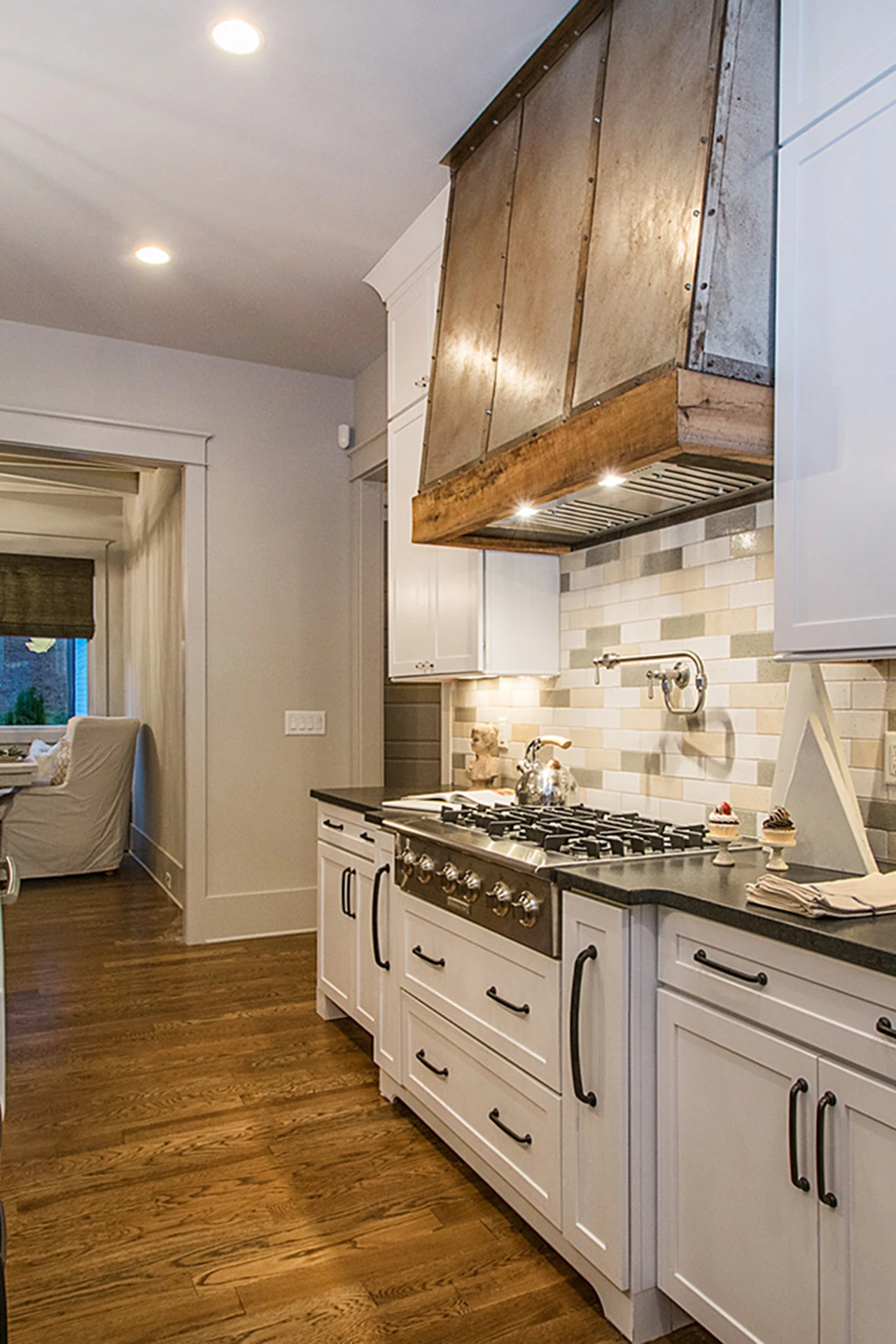 Galley kitchen with dark metal range hood, tile backsplash, stainless cooktop, and a hallway to a sitting room