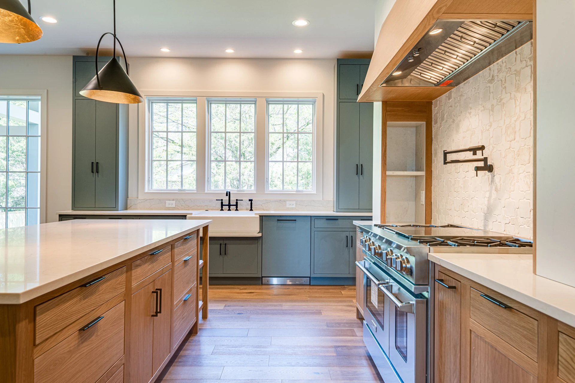 Kitchen with gray-green cabinets, wood island, farmhouse sink under three windows, stone backsplash, and wood range hood