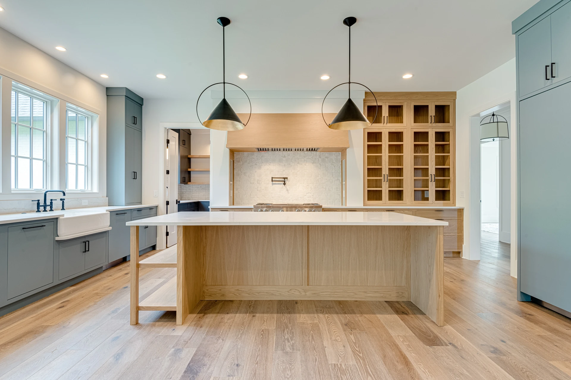 Kitchen with light blue cabinets, black dome pendants, white oak island, farmhouse sink, and glass-front pantry cabinets