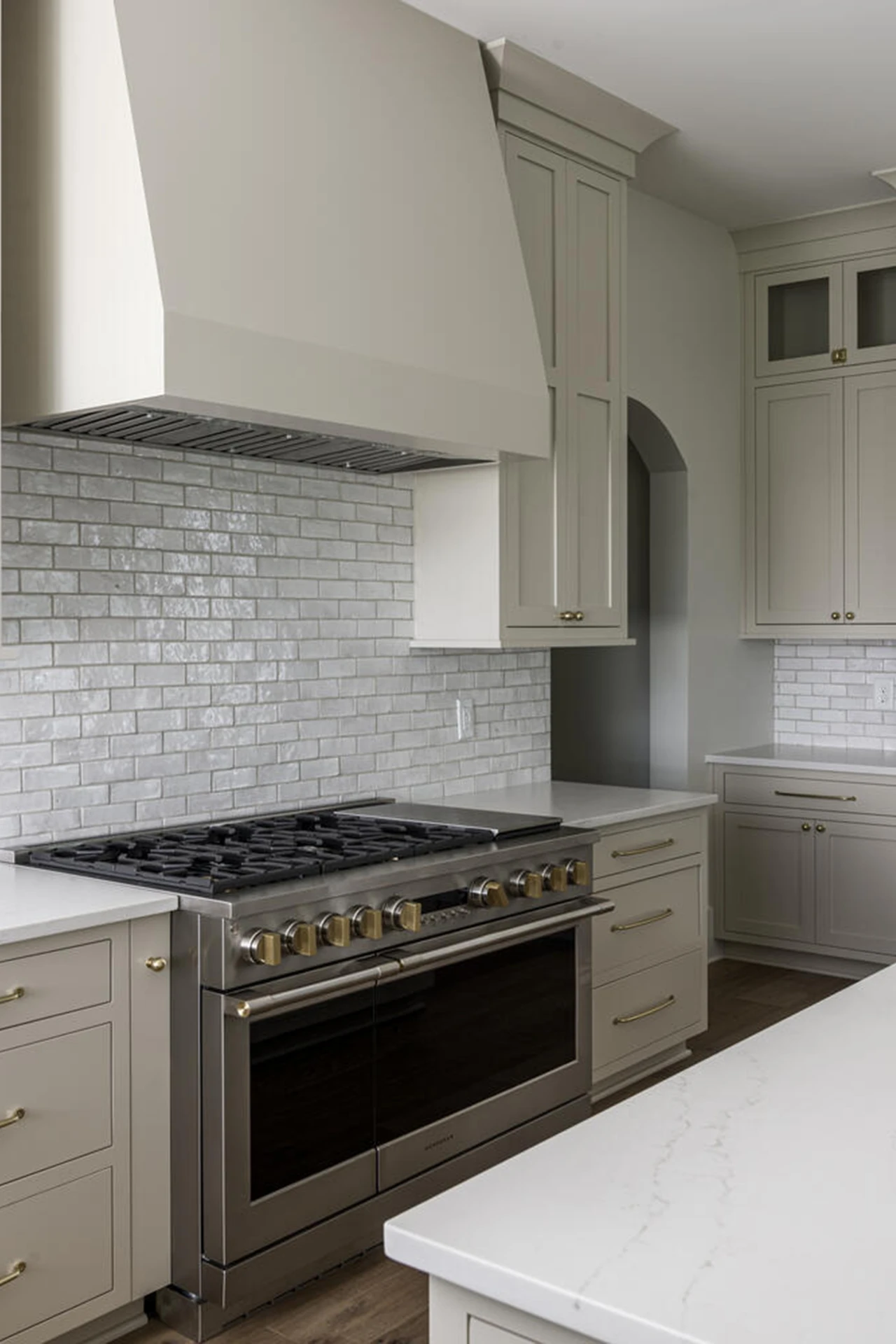 Greige kitchen with paneled range hood, gray subway backsplash, and a stainless professional range with brass knobs