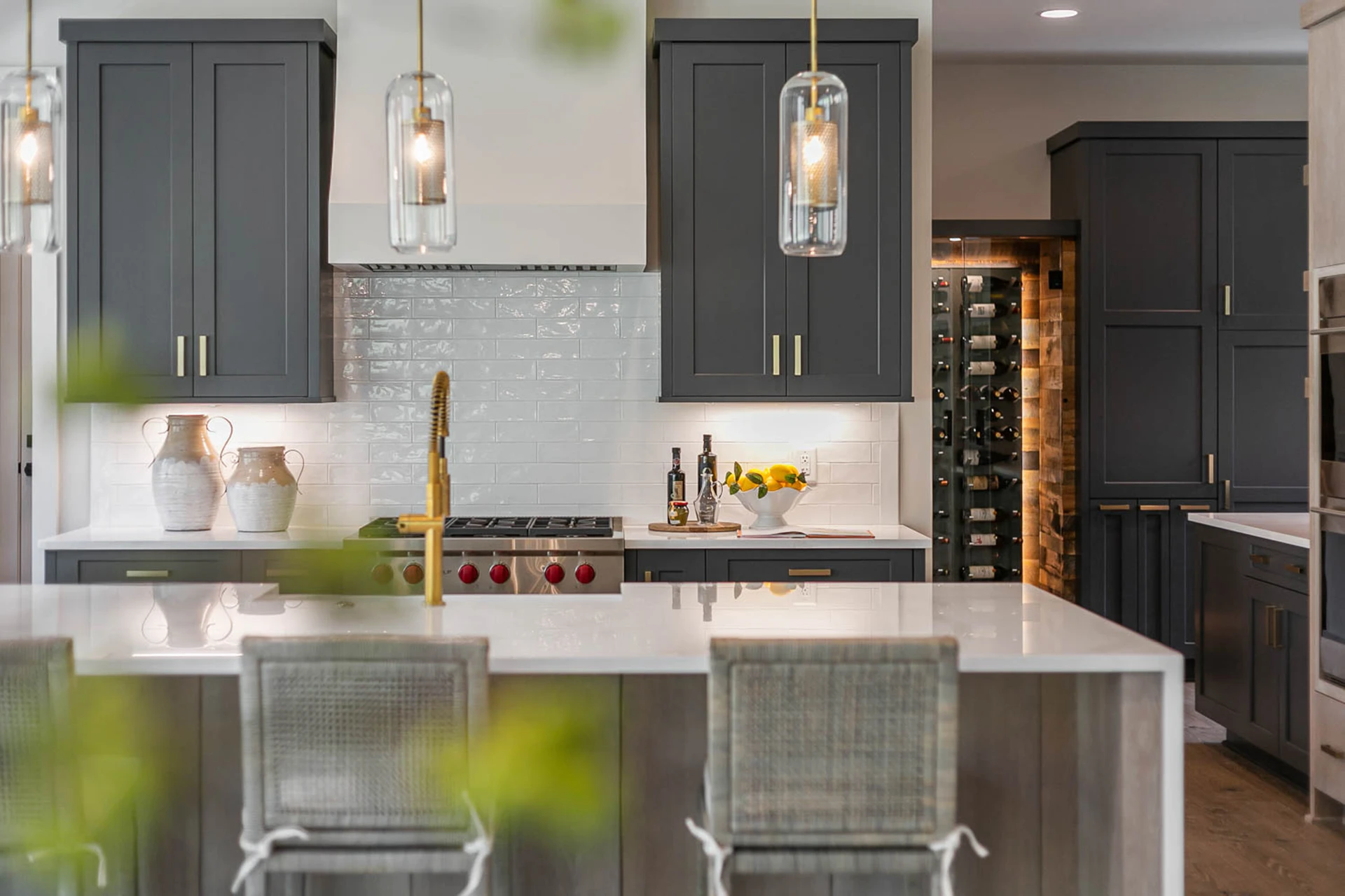 Dark gray kitchen with glass pendants, brass faucet, white brick backsplash, red-knob range, and a wine rack