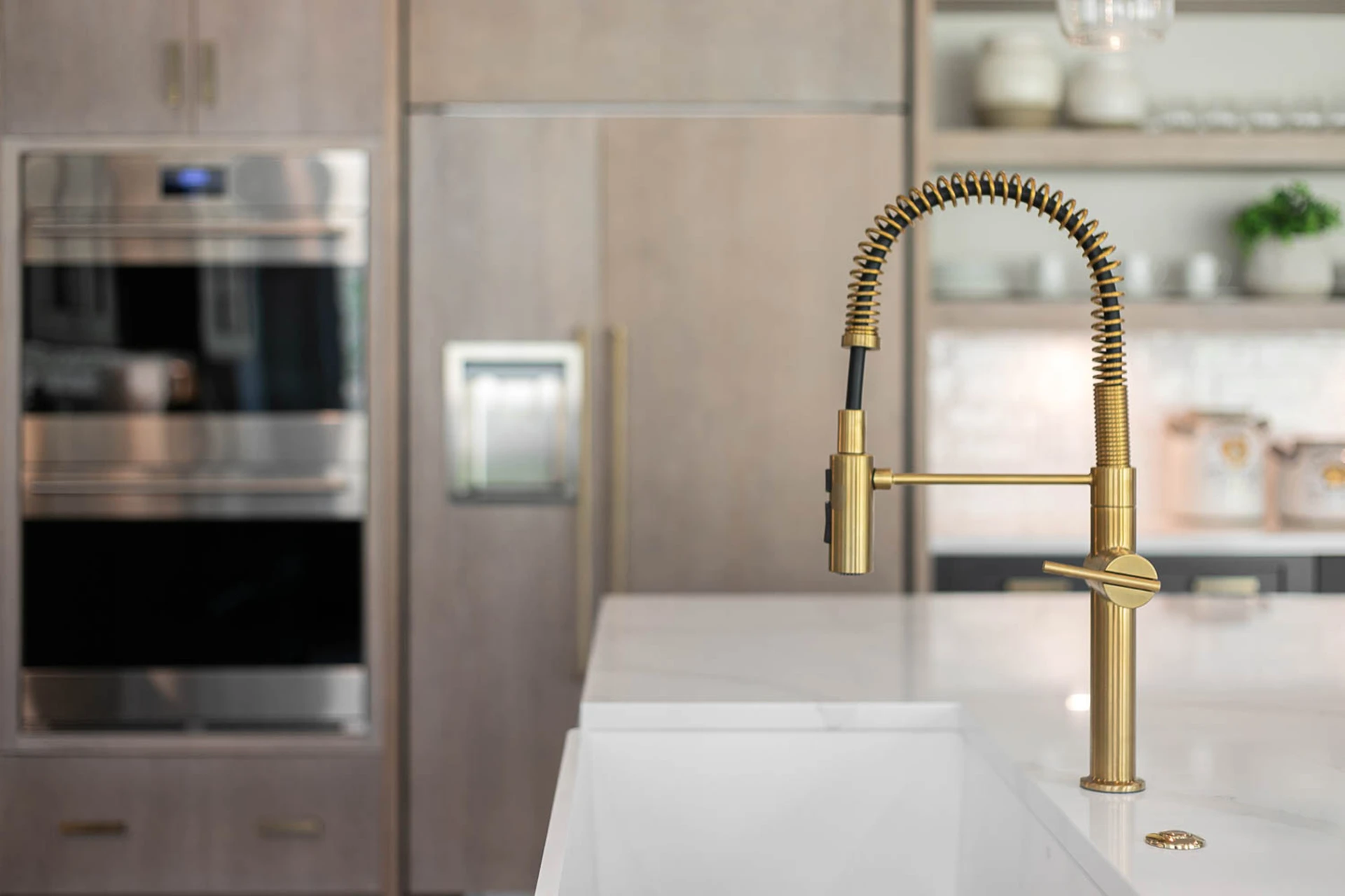 Close-up of a brushed brass spring-neck faucet over a white farmhouse sink, with stainless wall ovens behind