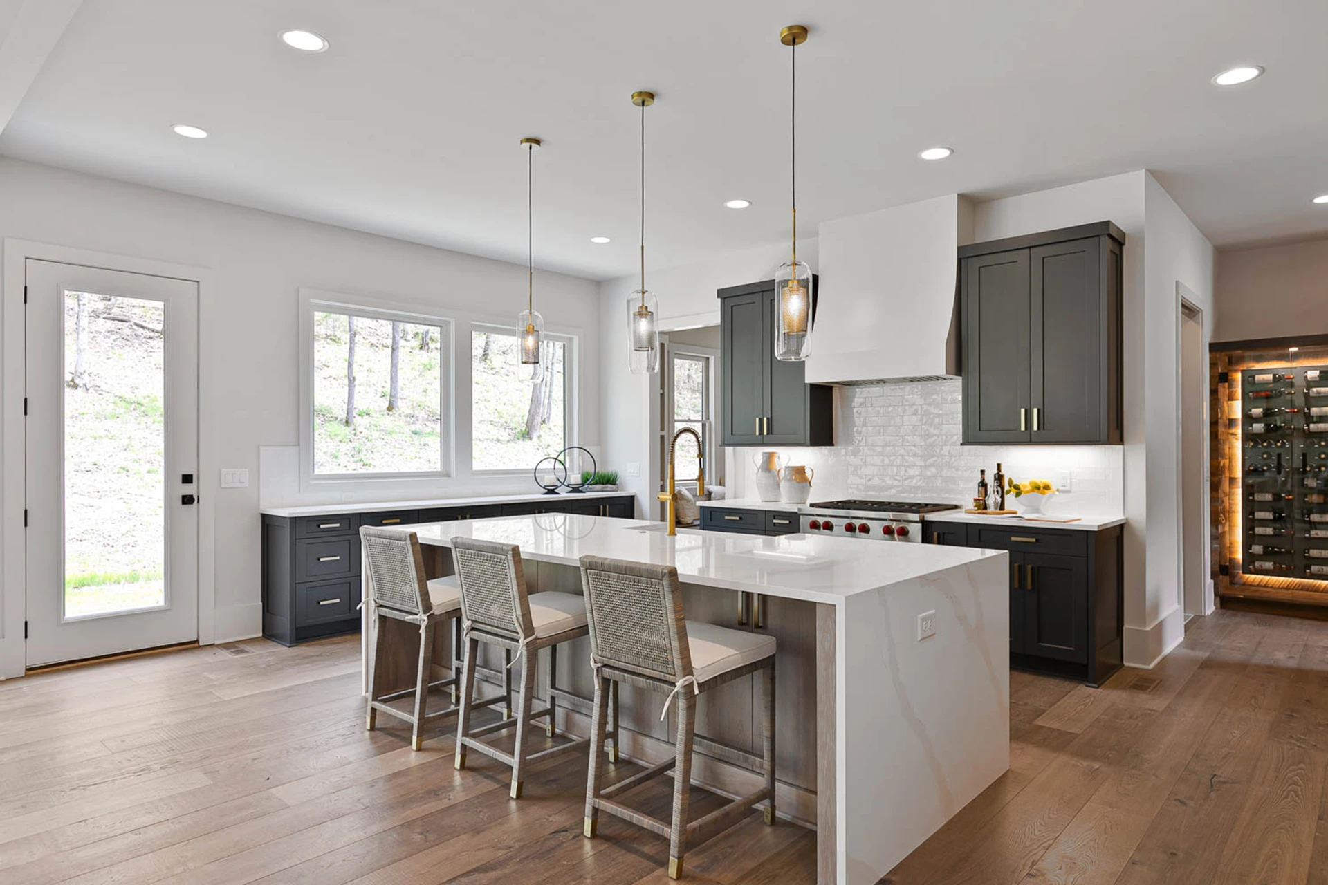 Kitchen with dark gray cabinets, white waterfall island, glass pendants, upholstered stools, and a wine display